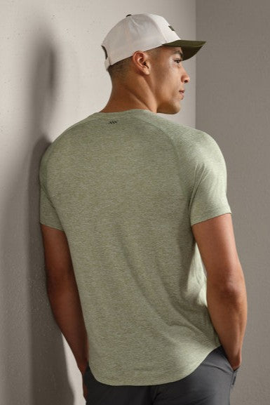 A man wearing the RHONE Atmosphere Tee in light green and a white and green baseball cap stands with his back to the camera, leaning against a light-colored wall.