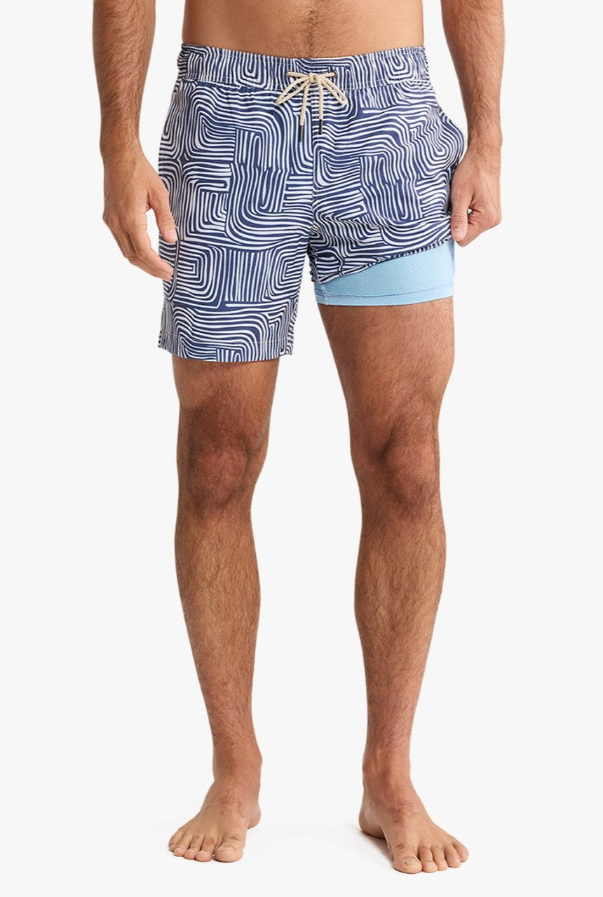 A man is wearing Fair Harbor Bayberry Trunk 7 Inch Wavy Sands shorts featuring blue and white patterns with a light blue panel, made from stretch fabric, standing barefoot against a plain white background.