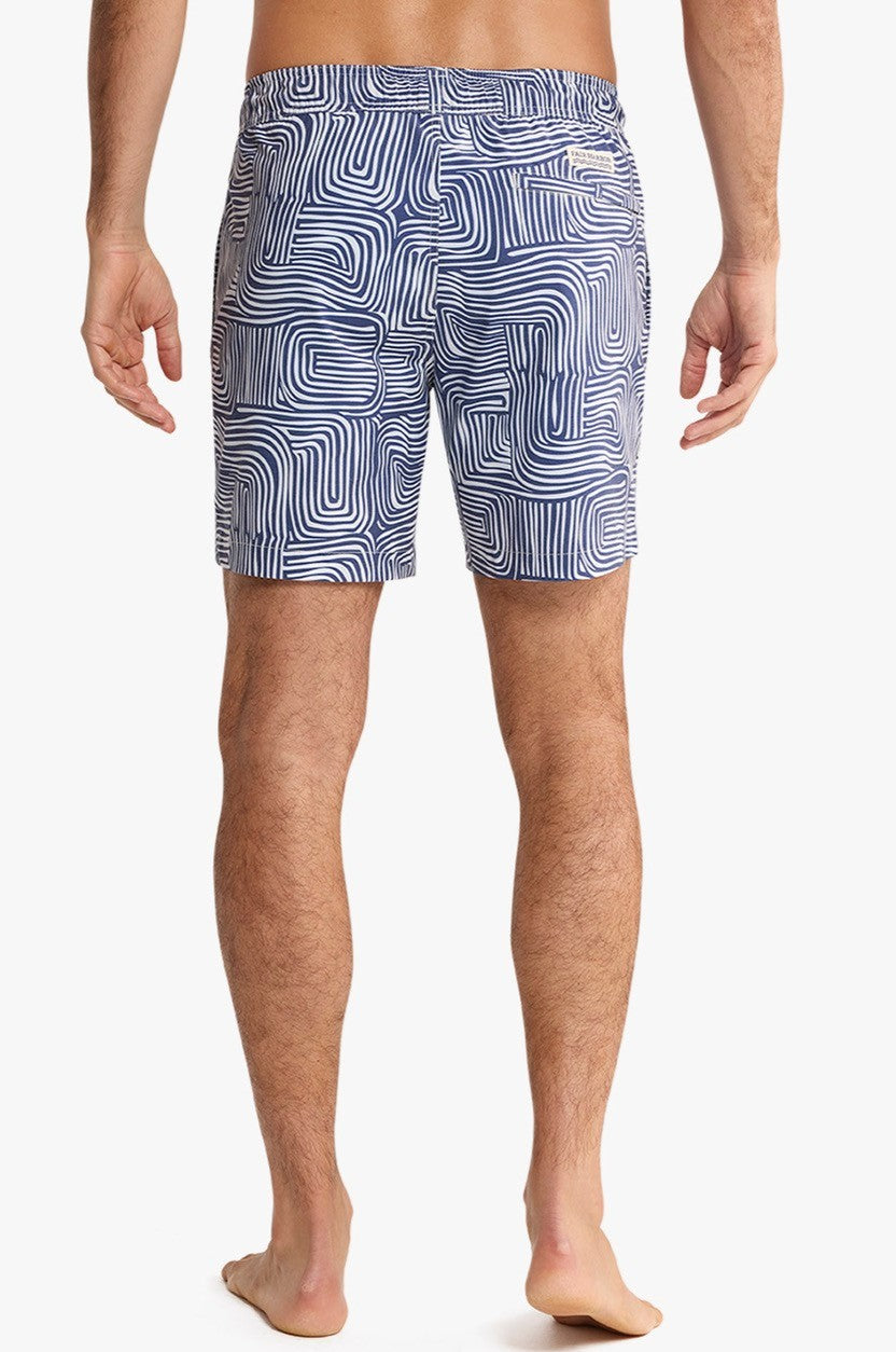 A barefoot person faces away in Fair Harbor Bayberry Trunk 7 Inch Wavy Sands swim shorts, featuring a blue and white abstract wavy line design and a BreezeKnit liner. The background is plain white.