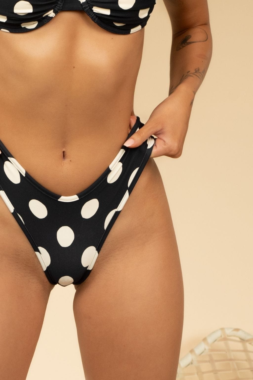 A person adjusts the Montce Polka Dot Lulu Bikini Bottom in Black Multi, featuring large white polka dots. Only their torso and upper legs are visible against a neutral beige background.