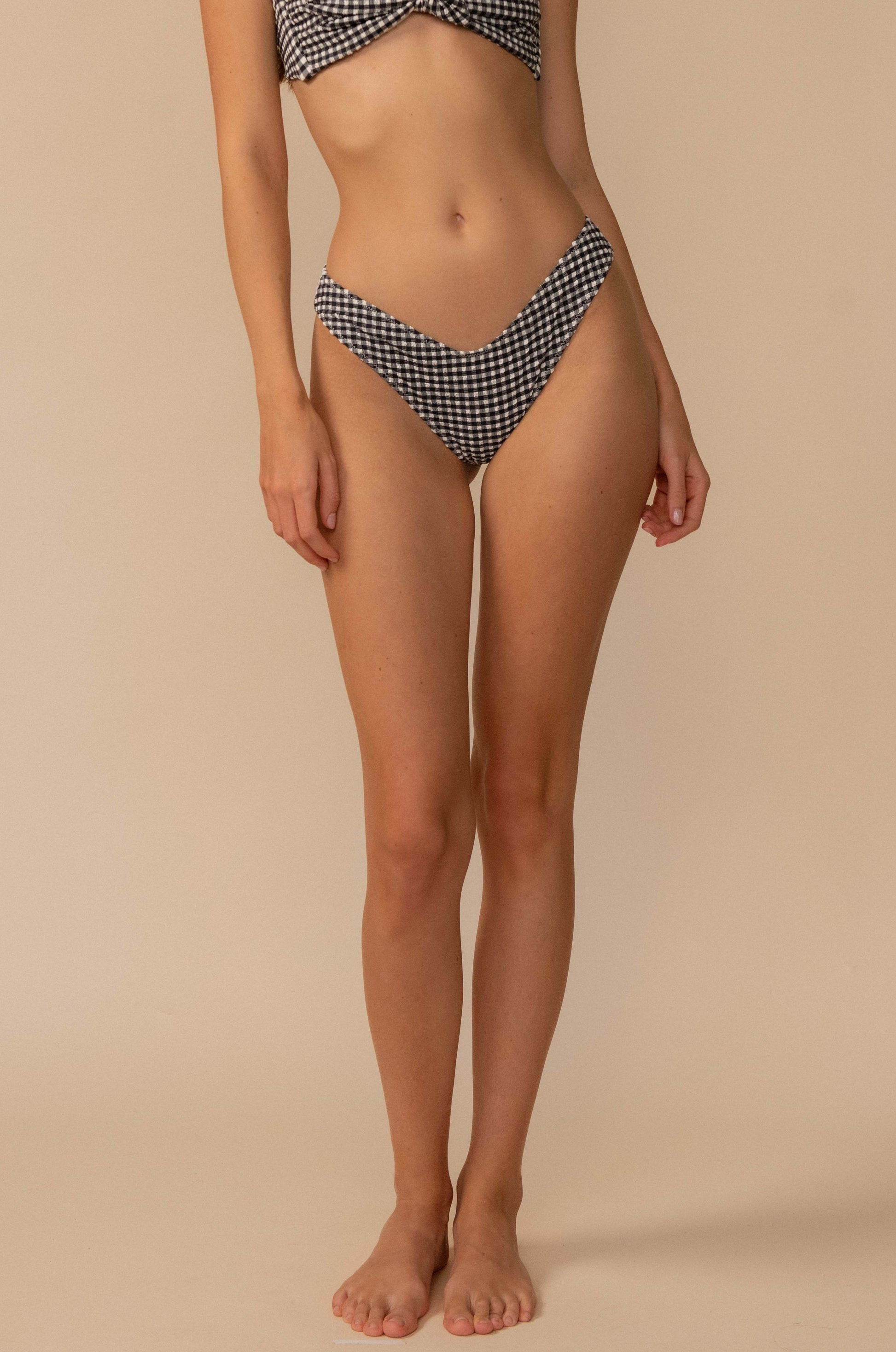 A woman stands barefoot against a beige background, wearing the Montce Black & White Gingham Lulu Bikini Bottom in Black Multi. Her face is not visible, and her arms rest relaxed at her sides.