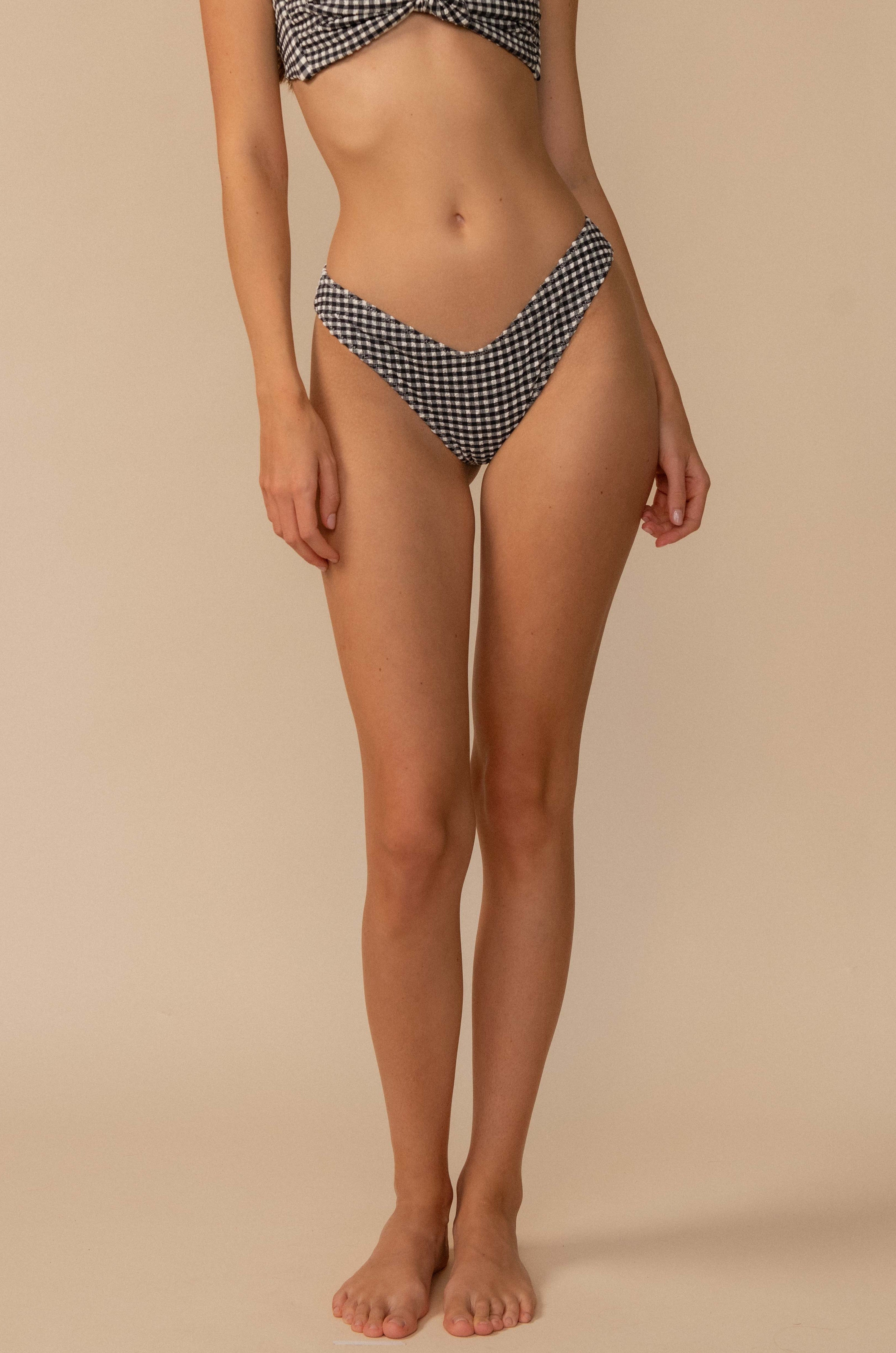 A woman stands barefoot against a beige background, wearing the Montce Black & White Gingham Lulu Bikini Bottom in Black Multi. Her face is not visible, and her arms rest relaxed at her sides.
