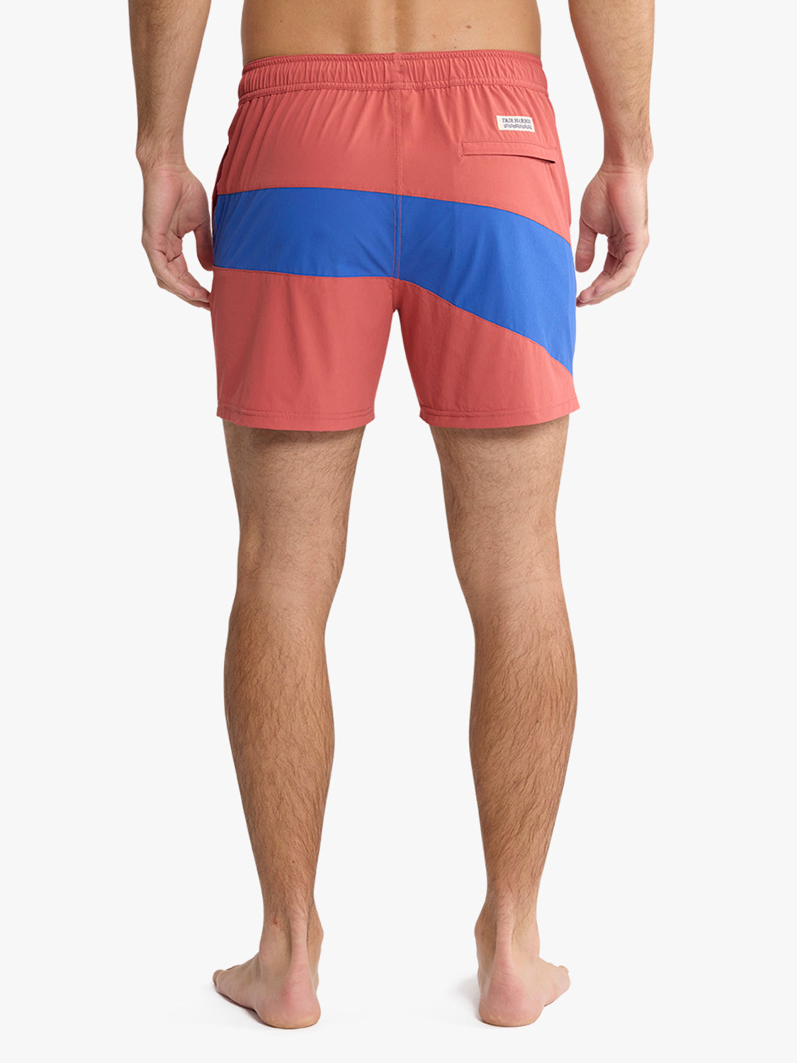 A man stands barefoot facing away, wearing Fair Harbor's Bungalow Trunk 5" in Nautical Colorblock—salmon quick-dry swim trunks with a diagonal blue stripe and small back pocket—against a plain white background.