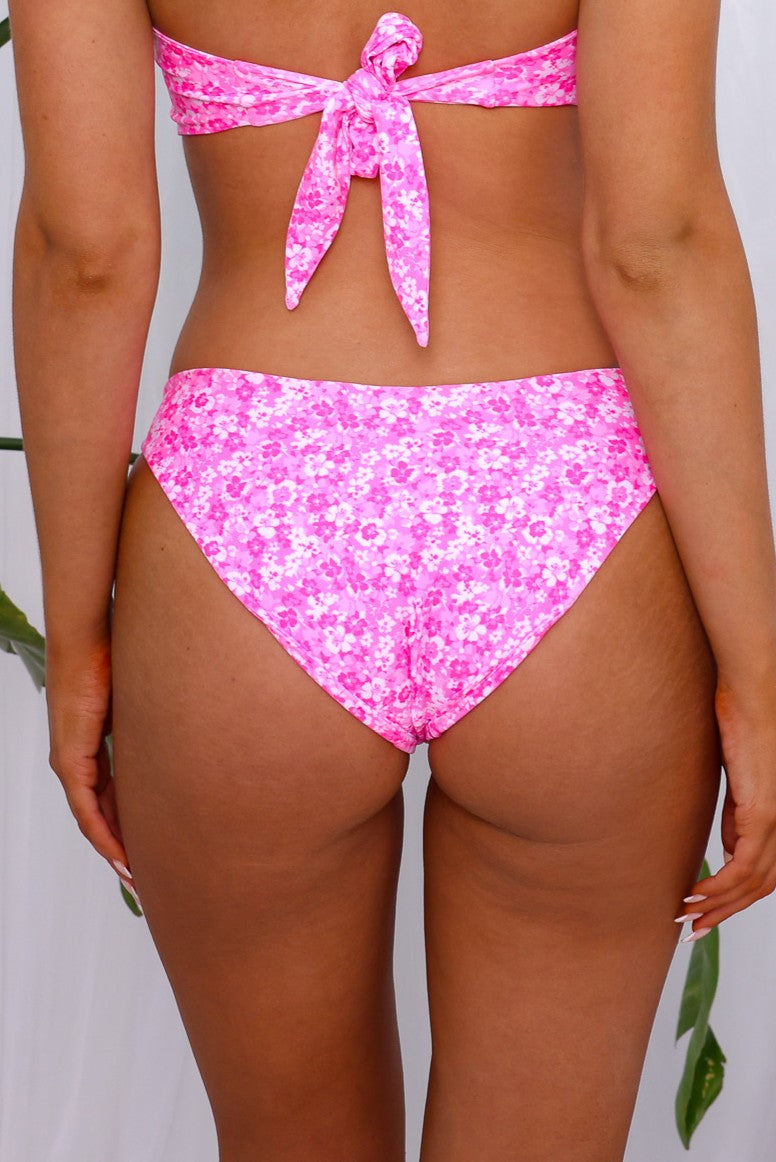 A person stands with their back to the camera, wearing the Berry Burst Minimal Full Coverage Bikini by Kulani Kinis—a pink floral set with high-cut bottoms and a strapless tanning top. Eco-friendly recycled polyester shines amid green leaves in the background.