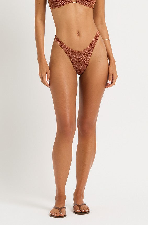 A woman is shown from shoulders to feet against a plain background, wearing the Bond-Eye Ginger Lurex Sign Brief bikini in orange and brown sandals, with her arms relaxed by her sides.