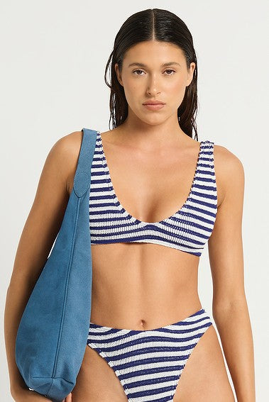 A woman with wet dark hair wears the Bond-Eye Navy White Scout Crop bikini, designed for medium to full support, and carries a large blue shoulder bag against a plain light background.