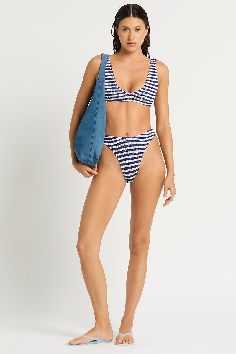 A woman with wet hair stands barefoot against a plain background, wearing the Bond-Eye Navy White Scout Crop, a blue and white striped bikini top offering medium to full support for larger busts. She holds a large blue shoulder bag and looks at the camera.