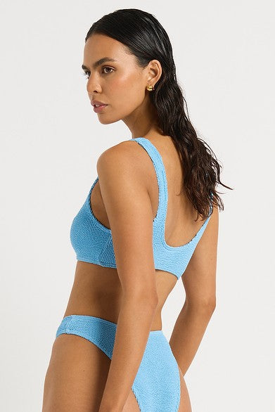 A woman with long dark hair stands sideways, glancing over her shoulder, wearing the Bond-Eye Cloud Blue Malibu 2.0 Crop bikini against a plain white background.