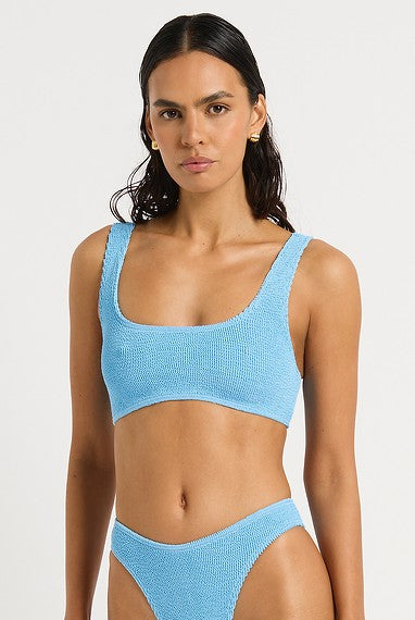 A woman with long dark hair wears the Bond-Eye Cloud Blue Malibu 2.0 Crop bikini set in light blue, featuring a square neckline and high-waisted bottoms, standing against a plain white background with a neutral expression.
