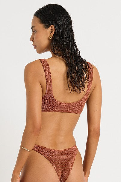 Wearing the Bond-Eye Ginger Lurex Maven Crop, a woman with wet, dark hair faces away. She pairs the rust-colored bikini set with gold hoop earrings and a thin bracelet against a plain white background.