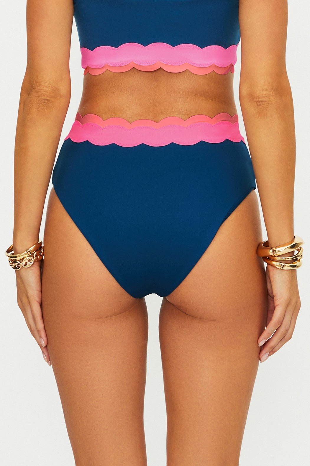 A person shown from the back wears the Beach Riot Seashell Isle Colorblock Harriet Bottom—a blue and pink high-waisted scalloped bikini—paired with gold and brown bracelets, set against a plain white background.