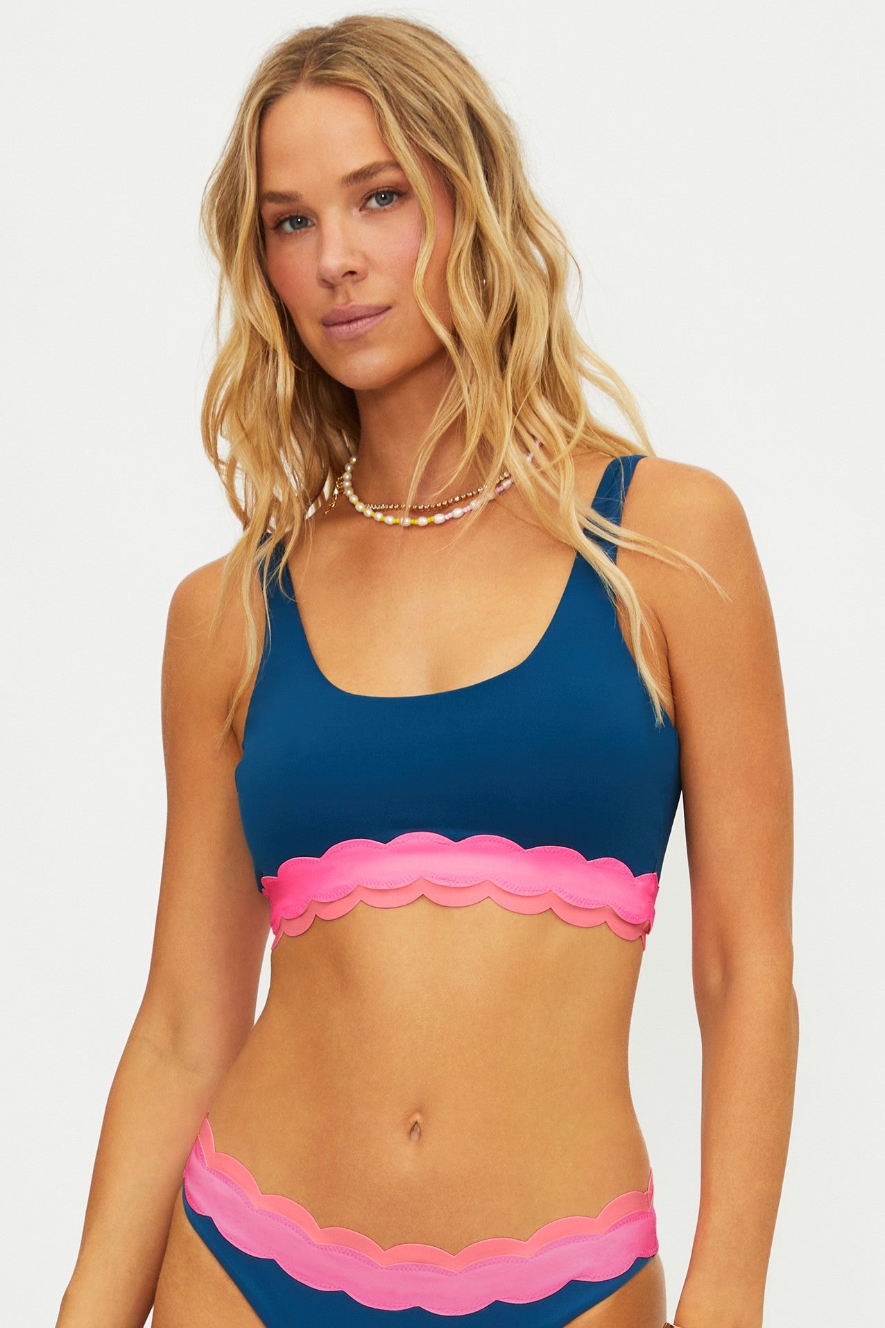 A woman with long wavy blonde hair wears the Beach Riot Seashell Isle Colorblock Marin Top, featuring a blue design with pink scallop hem and removable soft cups, paired with a beaded necklace against a plain white background.
