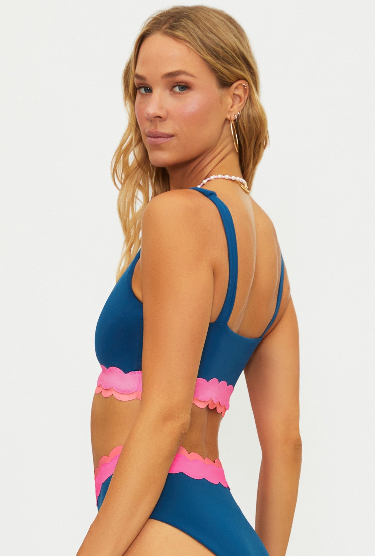 A woman with long blonde hair wears the Beach Riot Seashell Isle Colorblock Marin Top, featuring a blue and pink scallop hem, as she looks over her shoulder against a plain white background.
