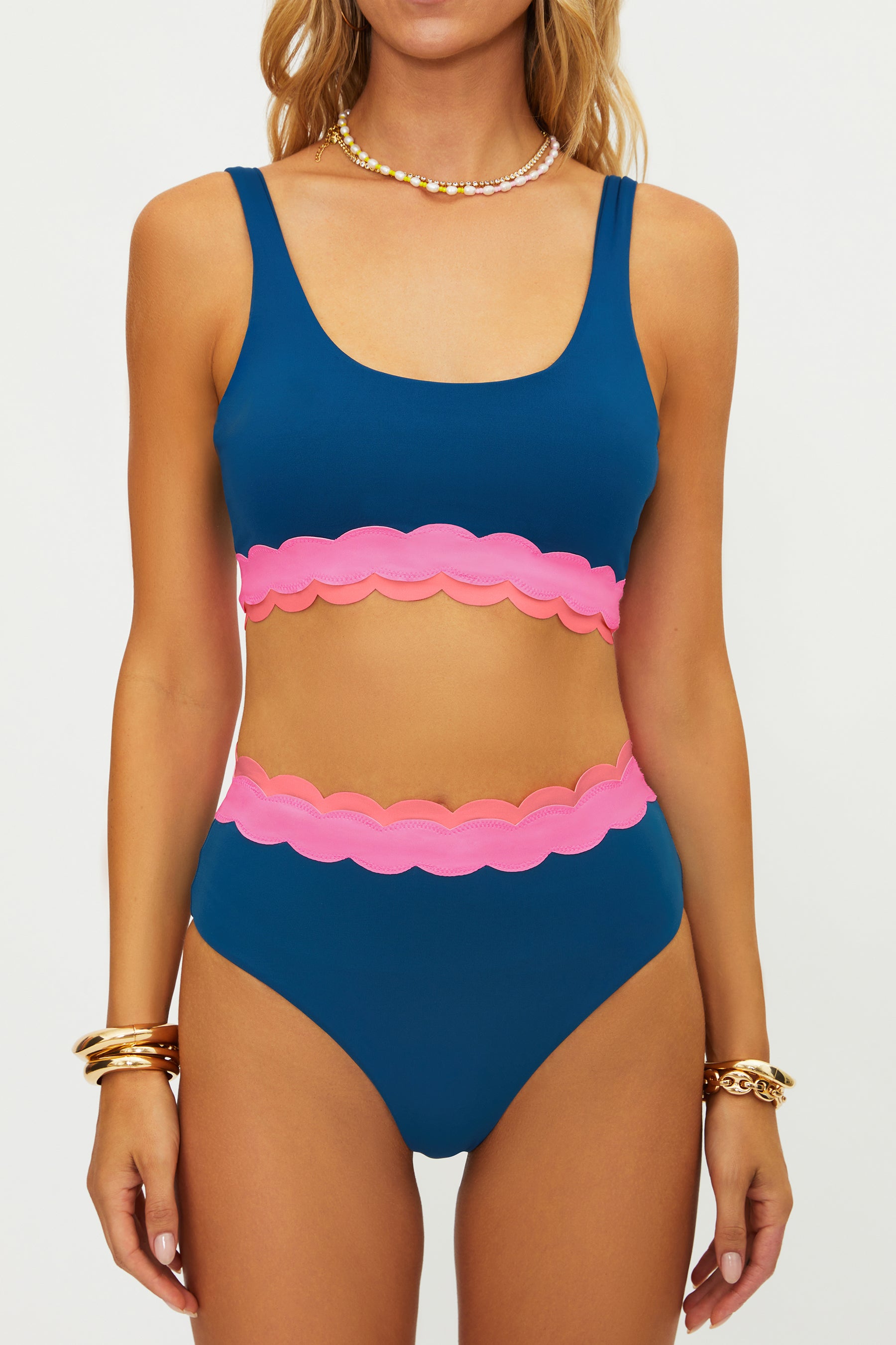 A woman wears the Beach Riot Seashell Isle Colorblock Marin Top, featuring a blue base with pink scallop trim on the chest and waist. She accessorizes with layered necklaces and gold bracelets. Her face is not shown.