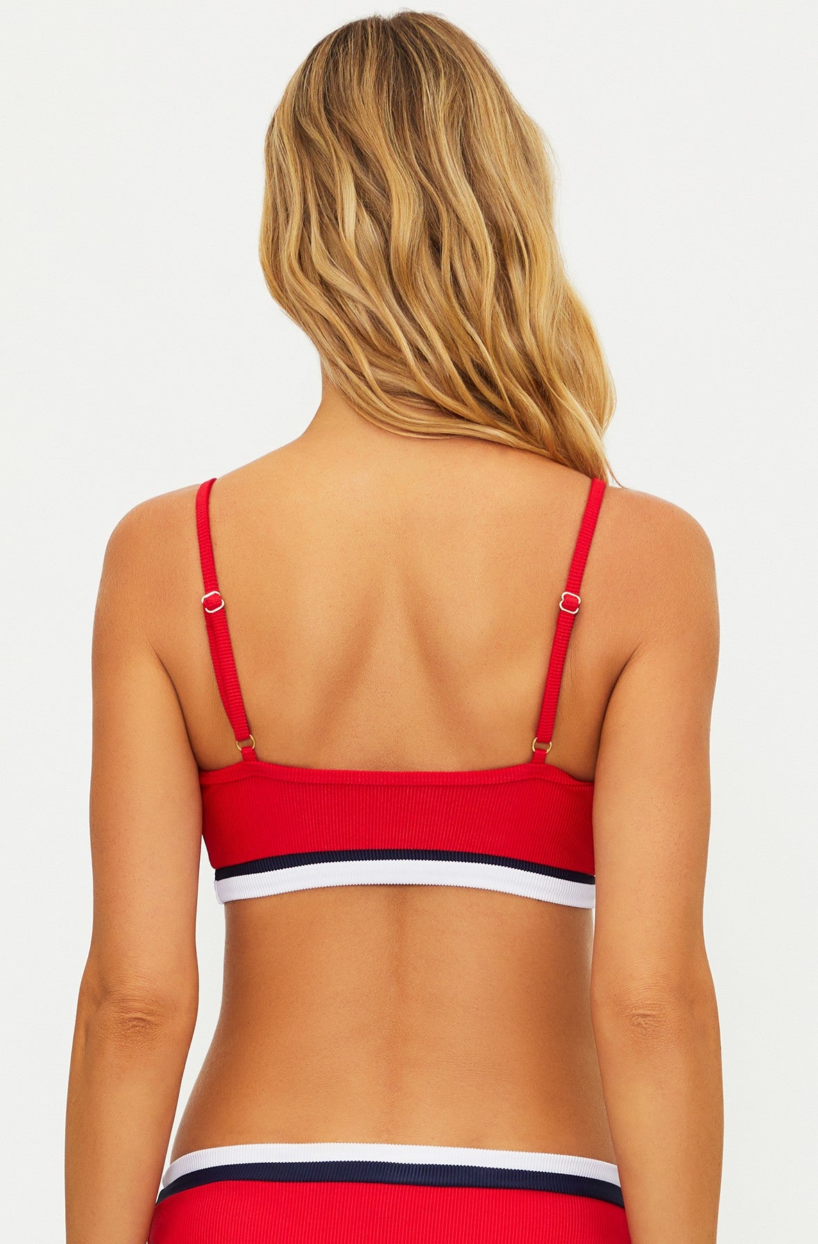 A woman with long, wavy blonde hair is seen from behind wearing the Beach Riot Crimson Firework Colorblock Val Bralette, featuring adjustable back straps and a white and black stripe on the bottom band.