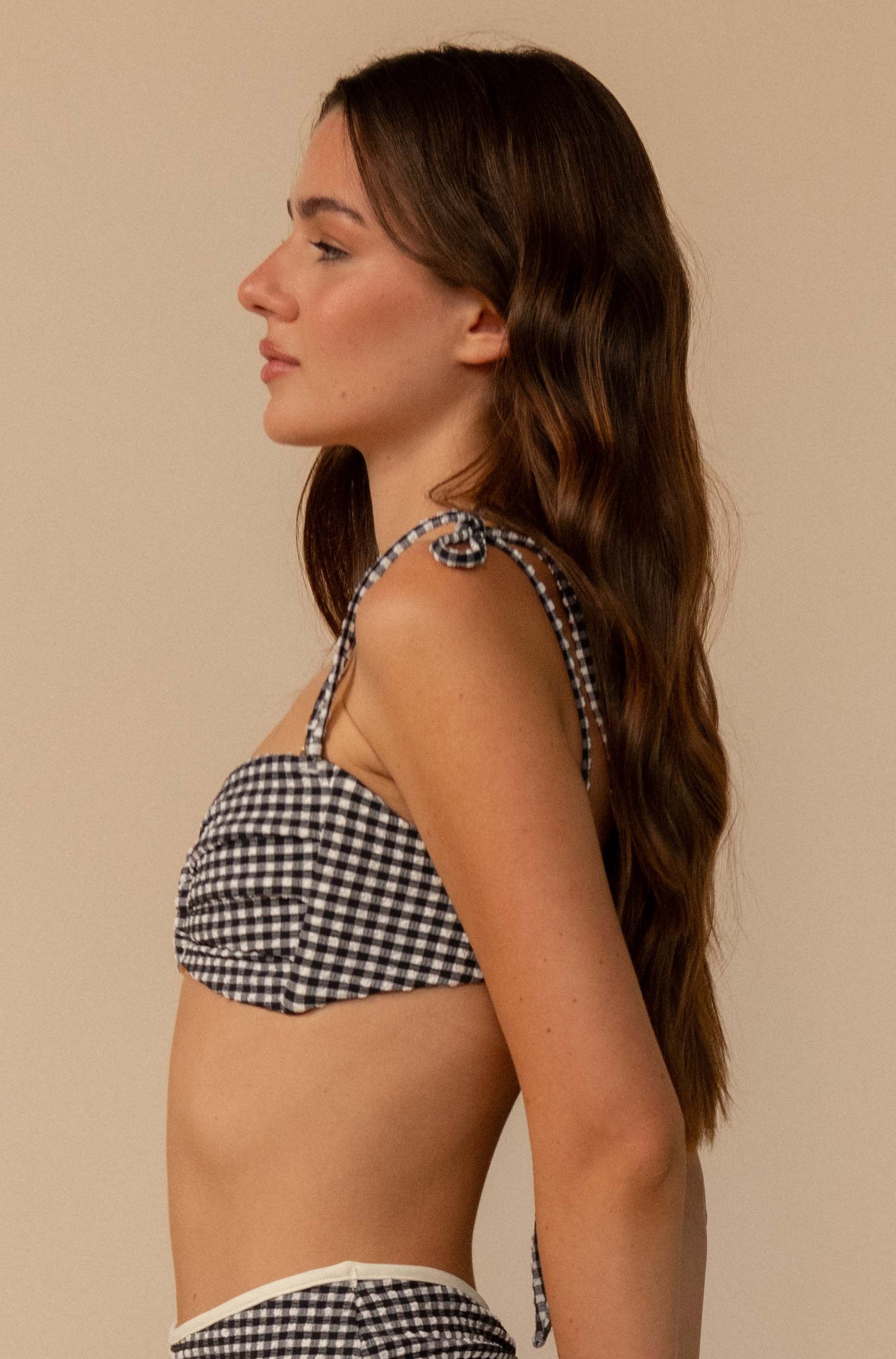 A woman with long brown hair stands in profile against a beige background, wearing the Montce Black & White Gingham Tori Ties Bandeau featuring knotted shoulder straps and ruched detailing.