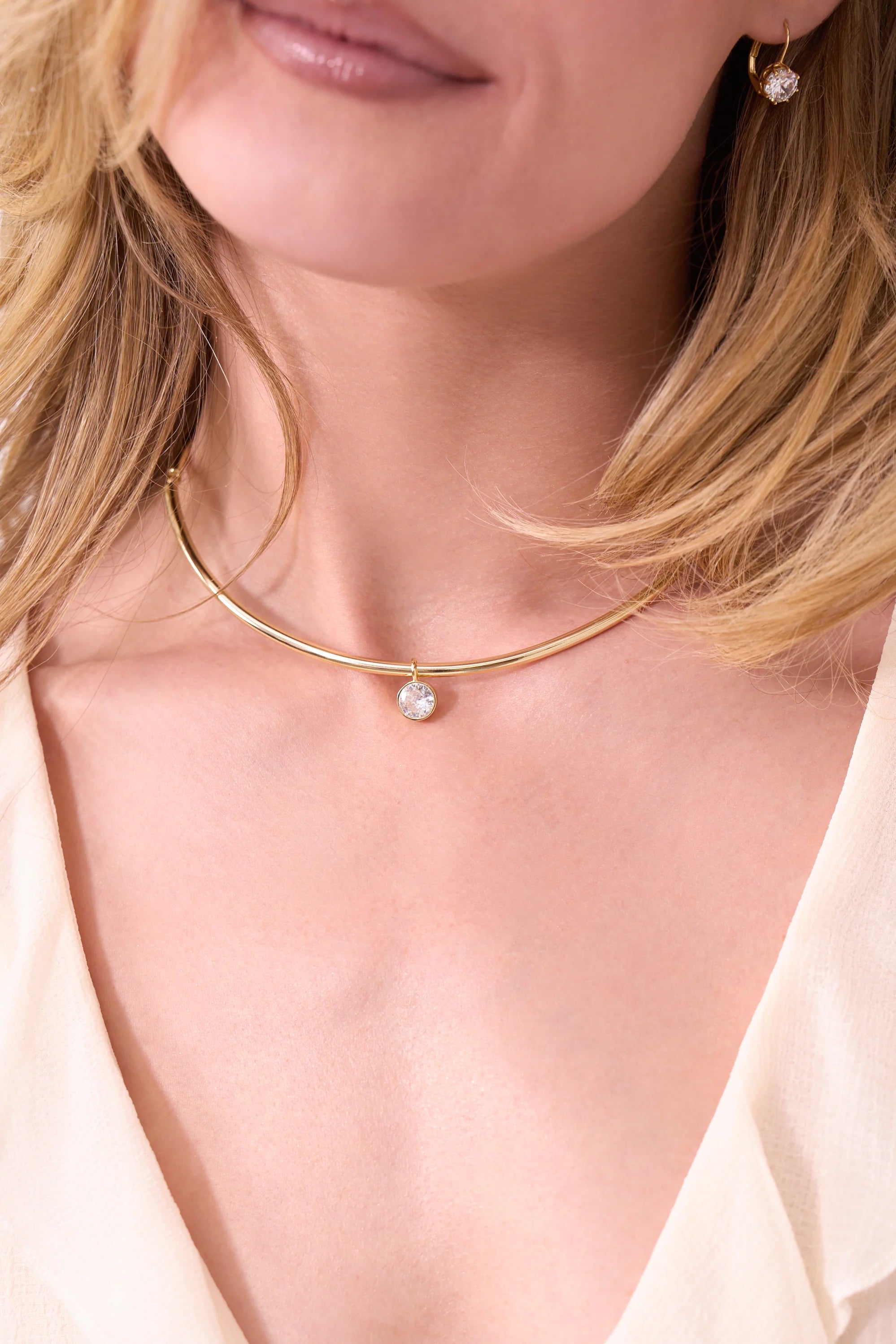 A blonde woman wears the Electric Picks Belle Necklace—a 14kt gold, 3mm thick chain with a single crystal pendant—paired with a small gold heart earring and a light V-neck top.