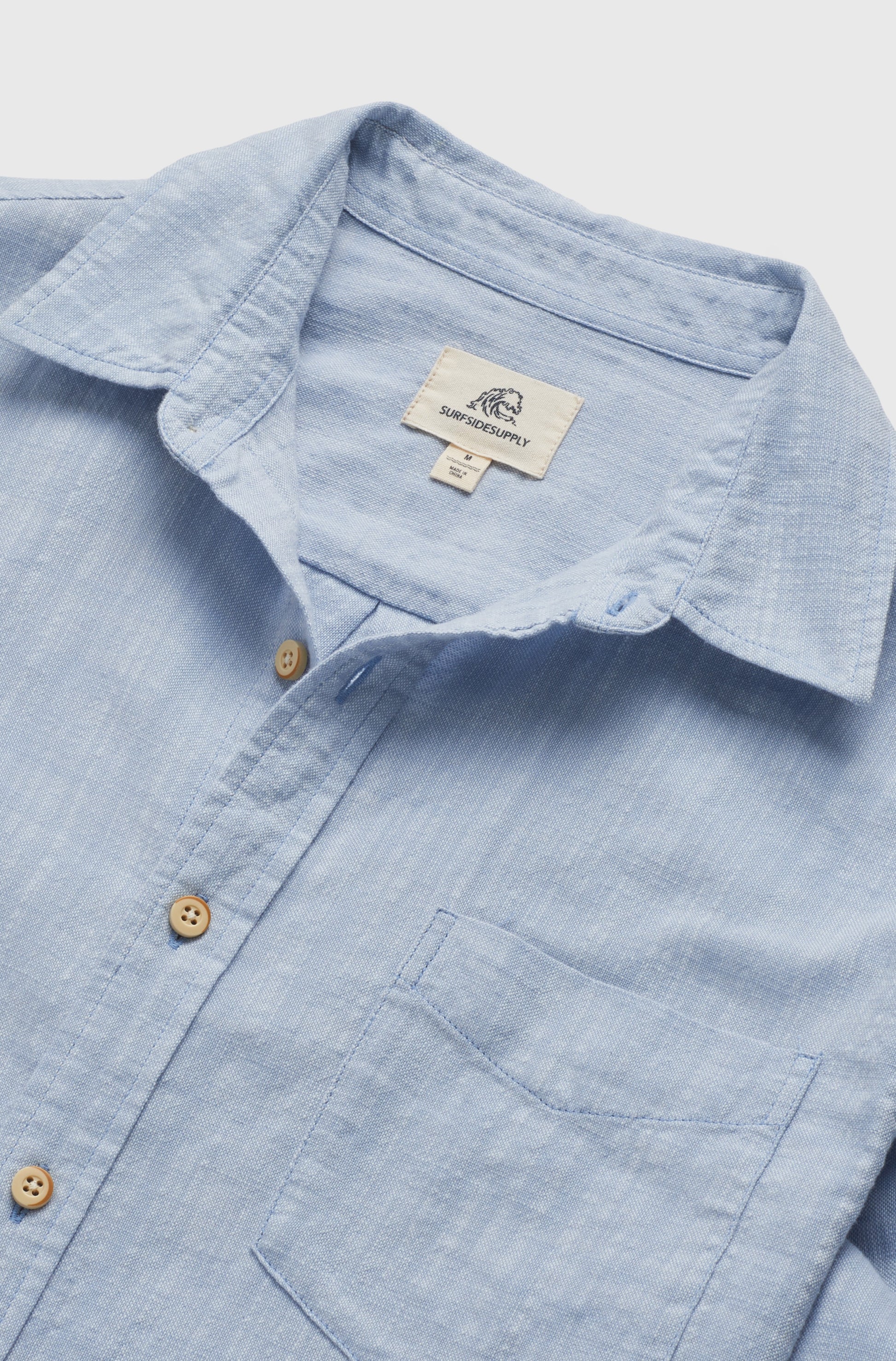 The Brian Long Sleeve 2tone Slub Shirt by Surfside Supply Co. is a light blue cotton shirt with a chest pocket, wooden buttons, and an inner collar label featuring the logo and text, shown laid flat on a white background.