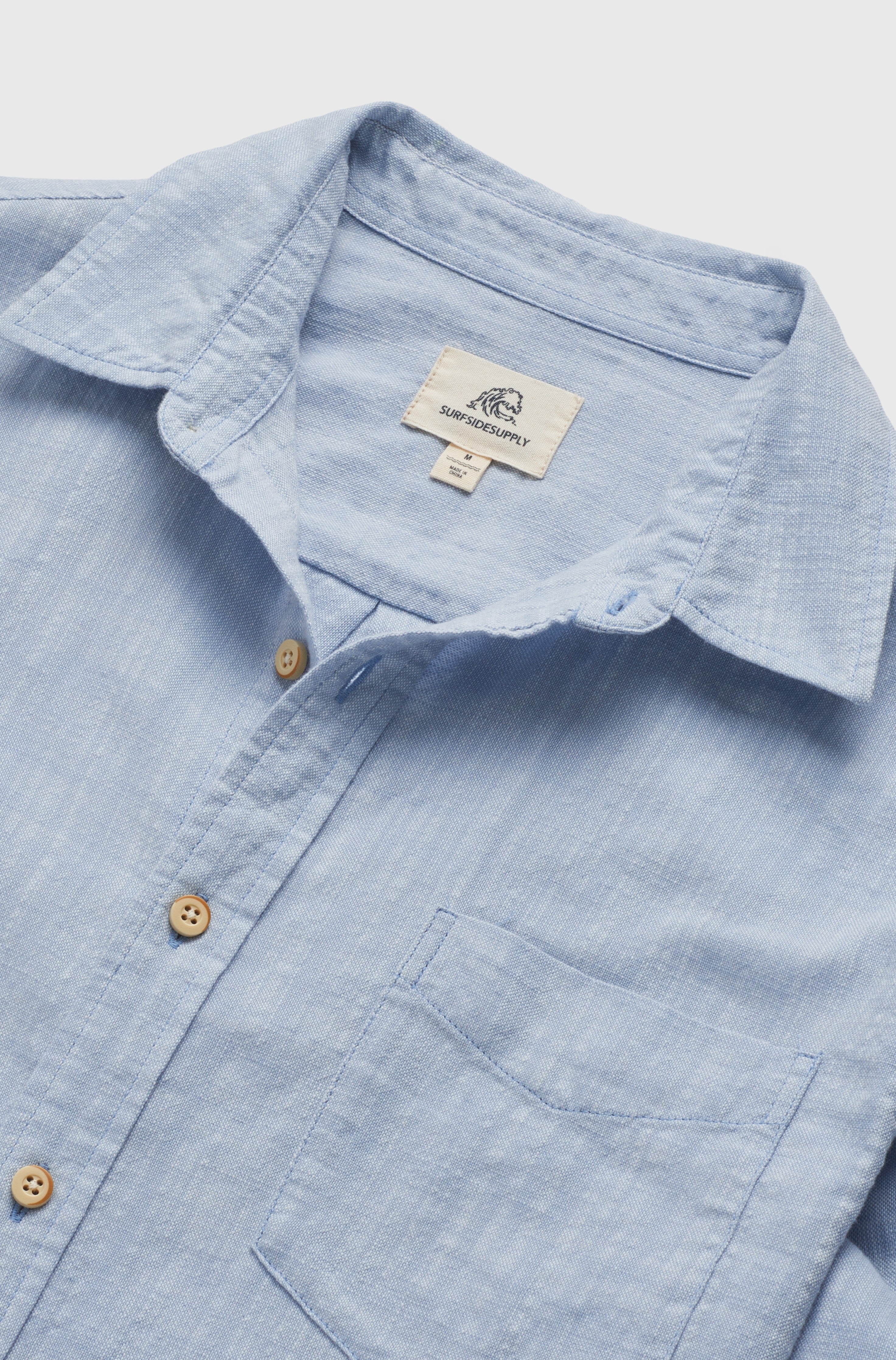 The Brian Long Sleeve 2tone Slub Shirt by Surfside Supply Co. is a light blue cotton shirt with a chest pocket, wooden buttons, and an inner collar label featuring the logo and text, shown laid flat on a white background.
