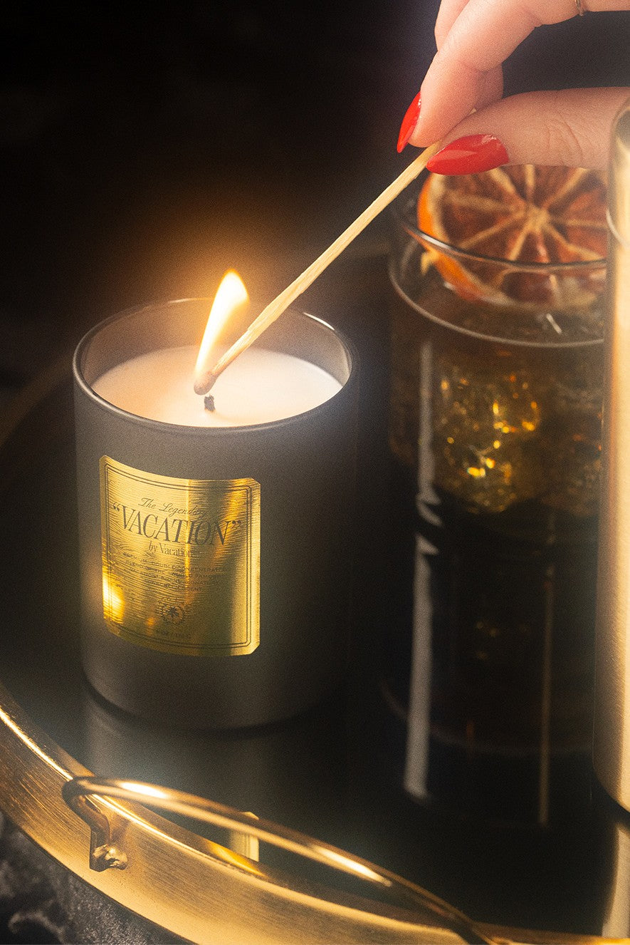 A hand with painted nails lights a Vacation By Vacation Candle by Vacation in a dark holder with a gold label on a shiny tray. Nearby, there’s a glass containing a drink and a slice of dried orange.