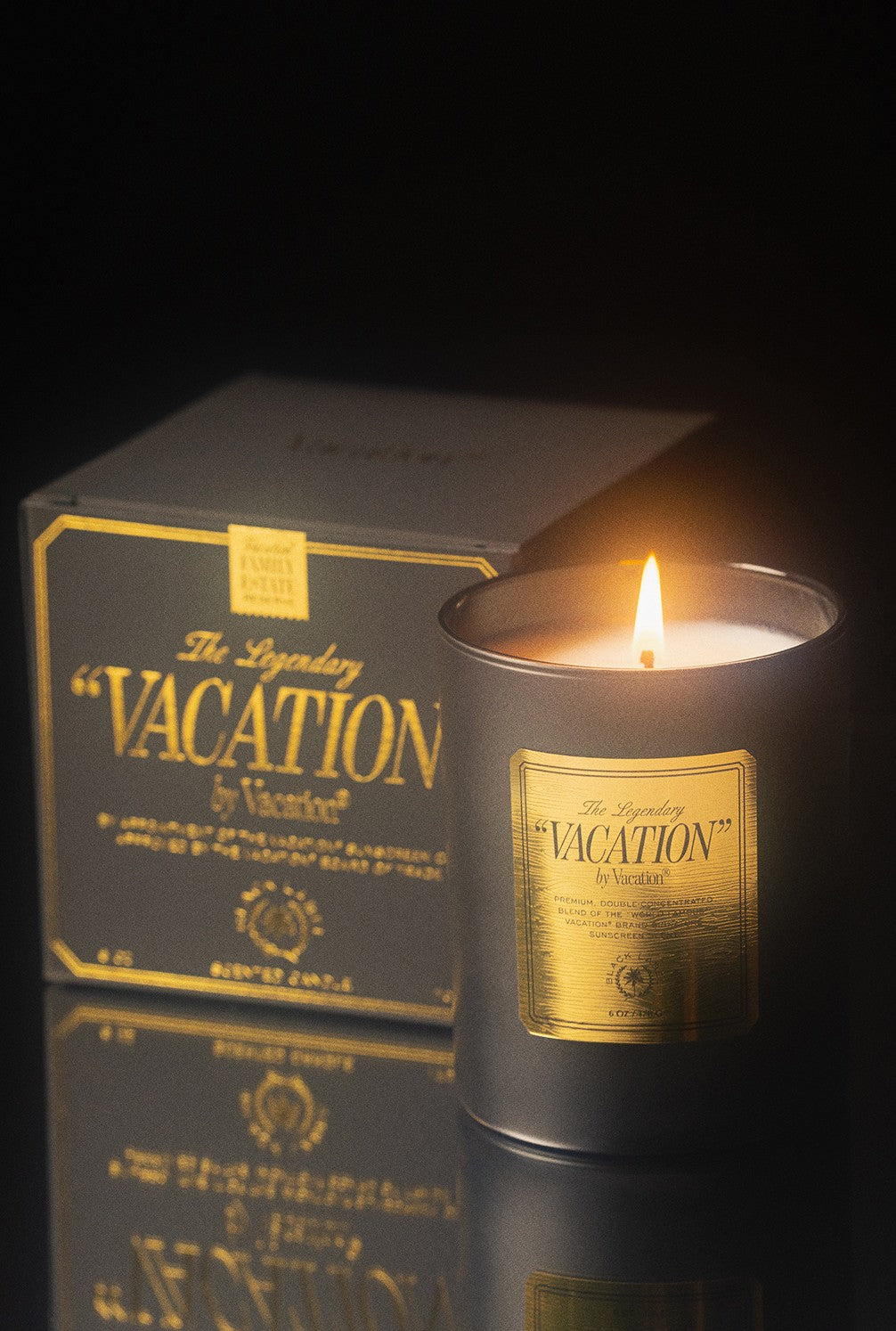 A Vacation By Vacation Candle by Vacation, in a gray glass holder labeled "The Legendary Vacation," sits with its matching box on a reflective surface against a dark background.
