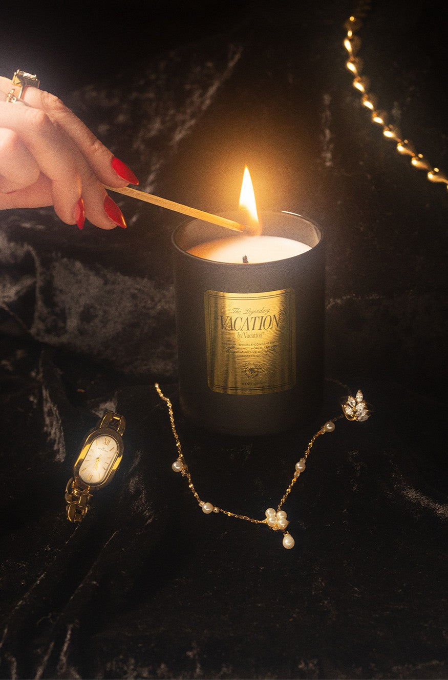 A hand with red nails lights the Vacation By Vacation Candle by Vacation with a match, while gold jewelry—a watch, necklace, ring, bracelet, and pearl earrings—rests elegantly on black velvet fabric.