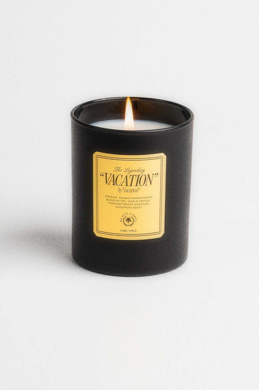 A lit Vacation By Vacation Candle by Vacation in a matte black jar with a yellow label is shown against a plain white background.