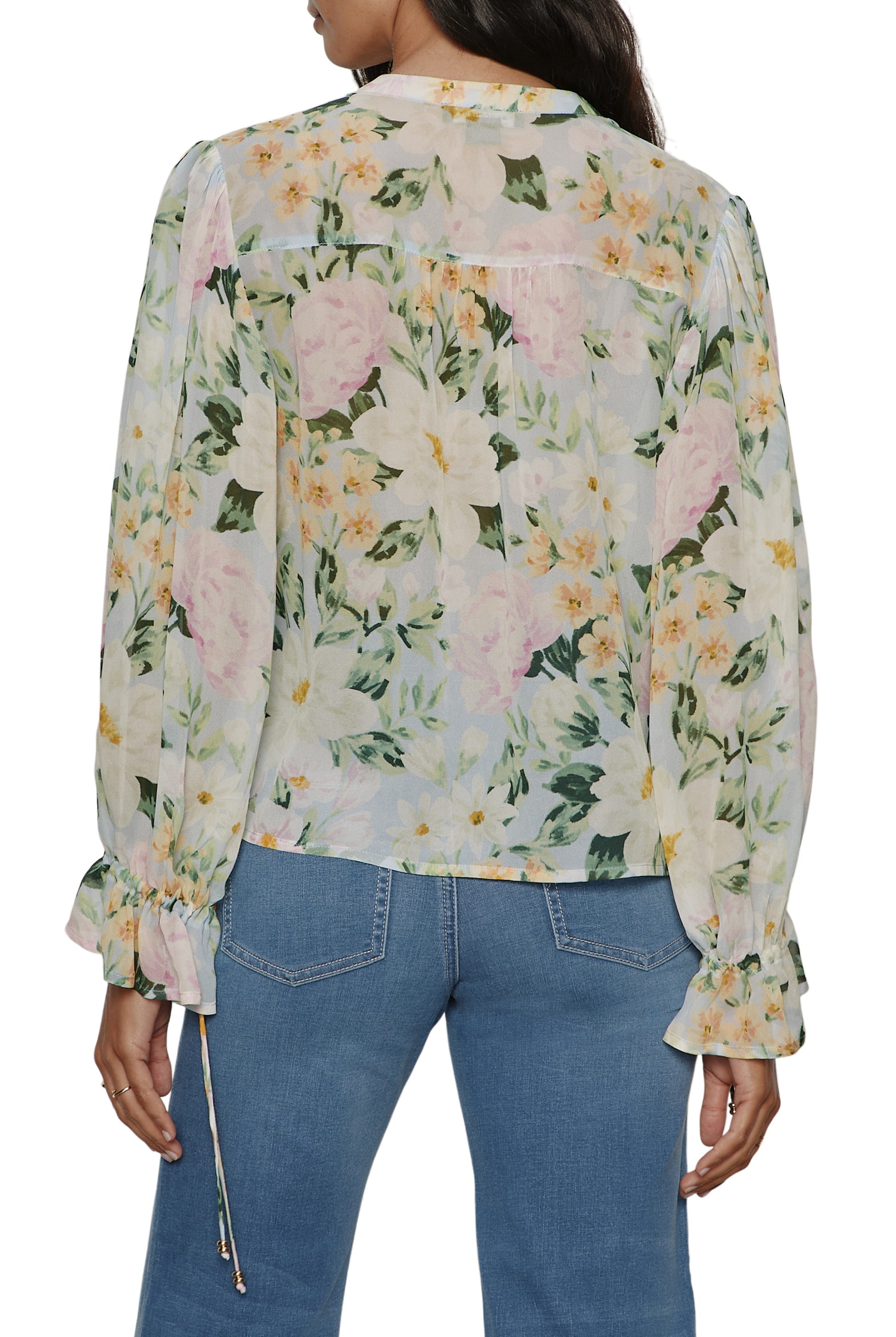 A person is seen from the back wearing the Sanctuary Femme Muse Button Blouse, a sheer, relaxed fit long sleeve top with tie cuffs and a floral pattern in pink, yellow, and white with green leaves, paired with light blue jeans.