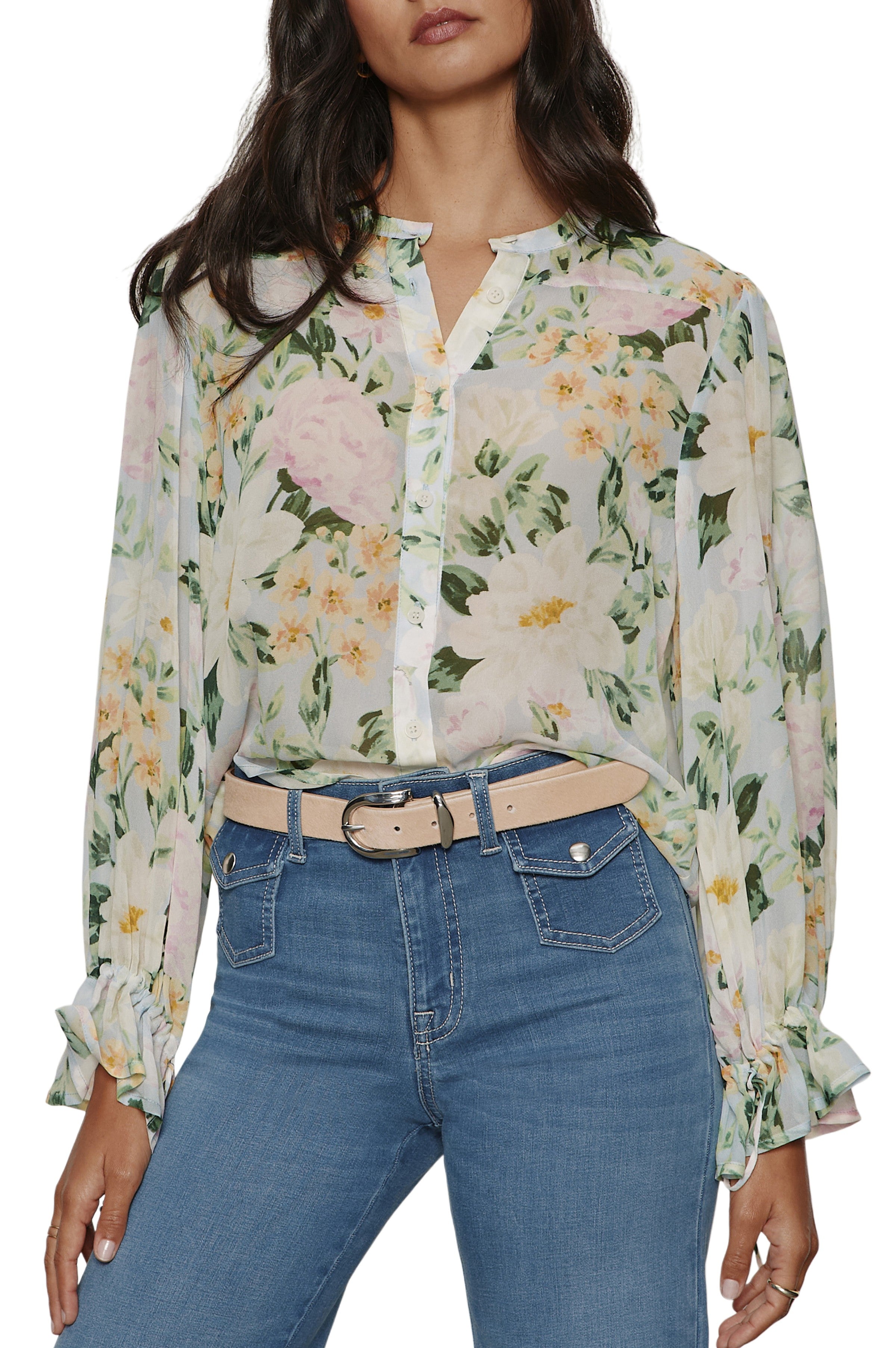 A woman wears the Femme Muse Button Blouse by Sanctuary—a relaxed fit, sheer long sleeve top with pastel florals, tie cuffs, and half-buttoned front—tucked into high-waisted blue jeans with a beige belt. Her head is partially cropped out of frame.
