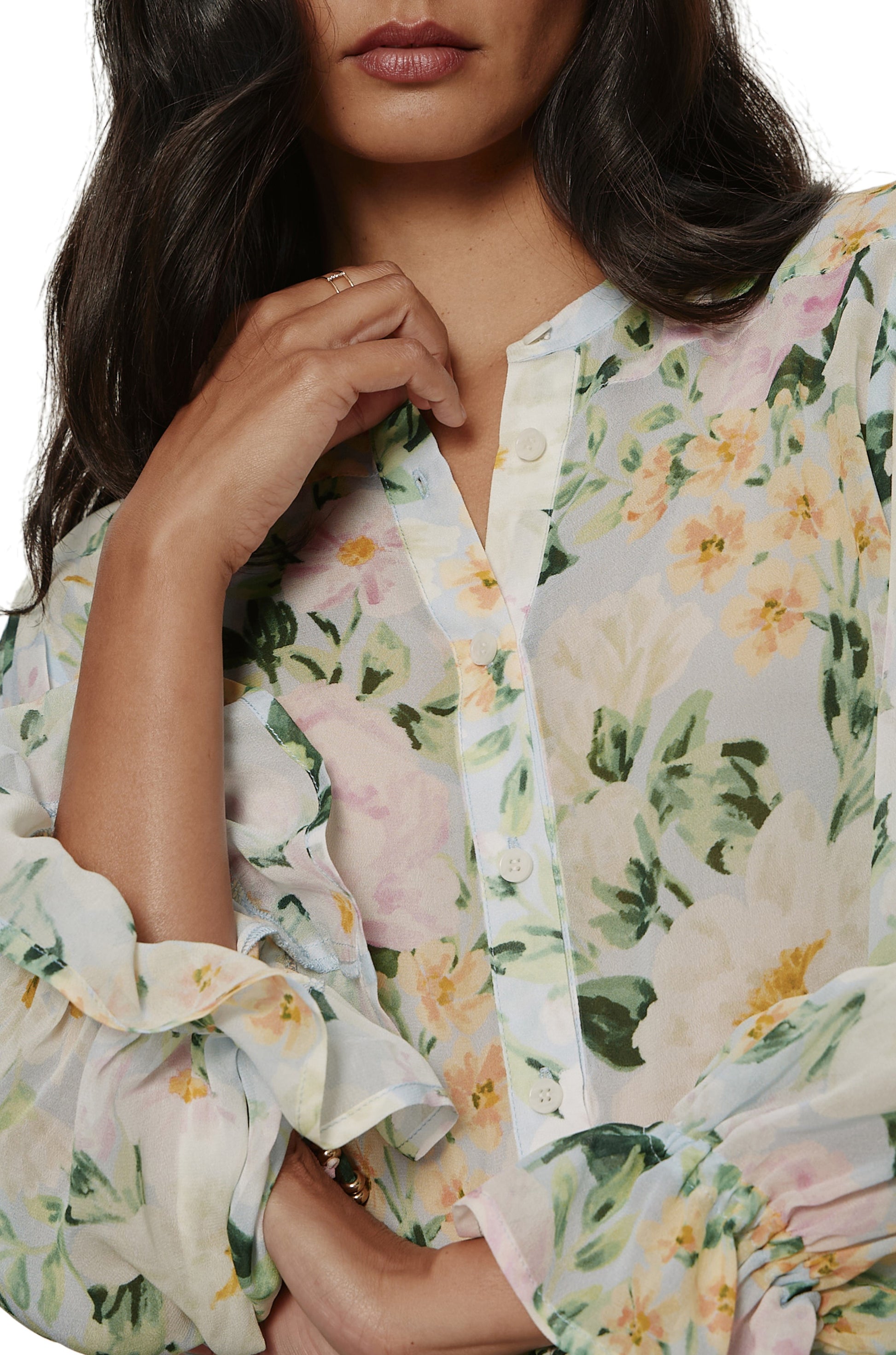 A woman poses in the Sanctuary Femme Muse Button Blouse, a pastel floral long-sleeve top with ruffled sleeves, tie cuffs, and sheer fabric. Its relaxed fit and soft details create a graceful look as her face is partially visible.