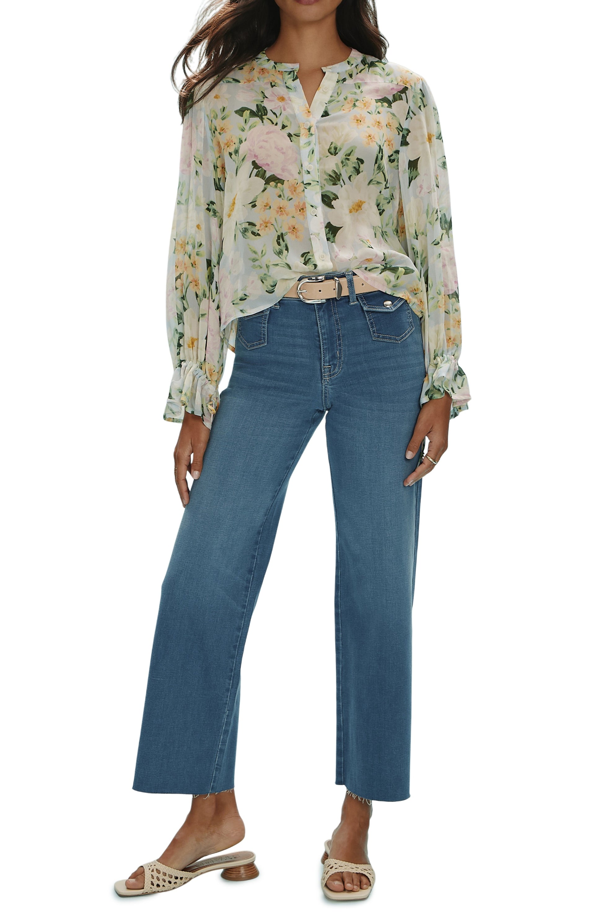 A woman wears the Femme Muse Button Blouse by Sanctuary, a light floral relaxed-fit long-sleeve top with tie cuffs, tucked into high-waisted wide-leg blue jeans and a beige belt. She finishes the look with tan slide sandals and a ring on her left hand.