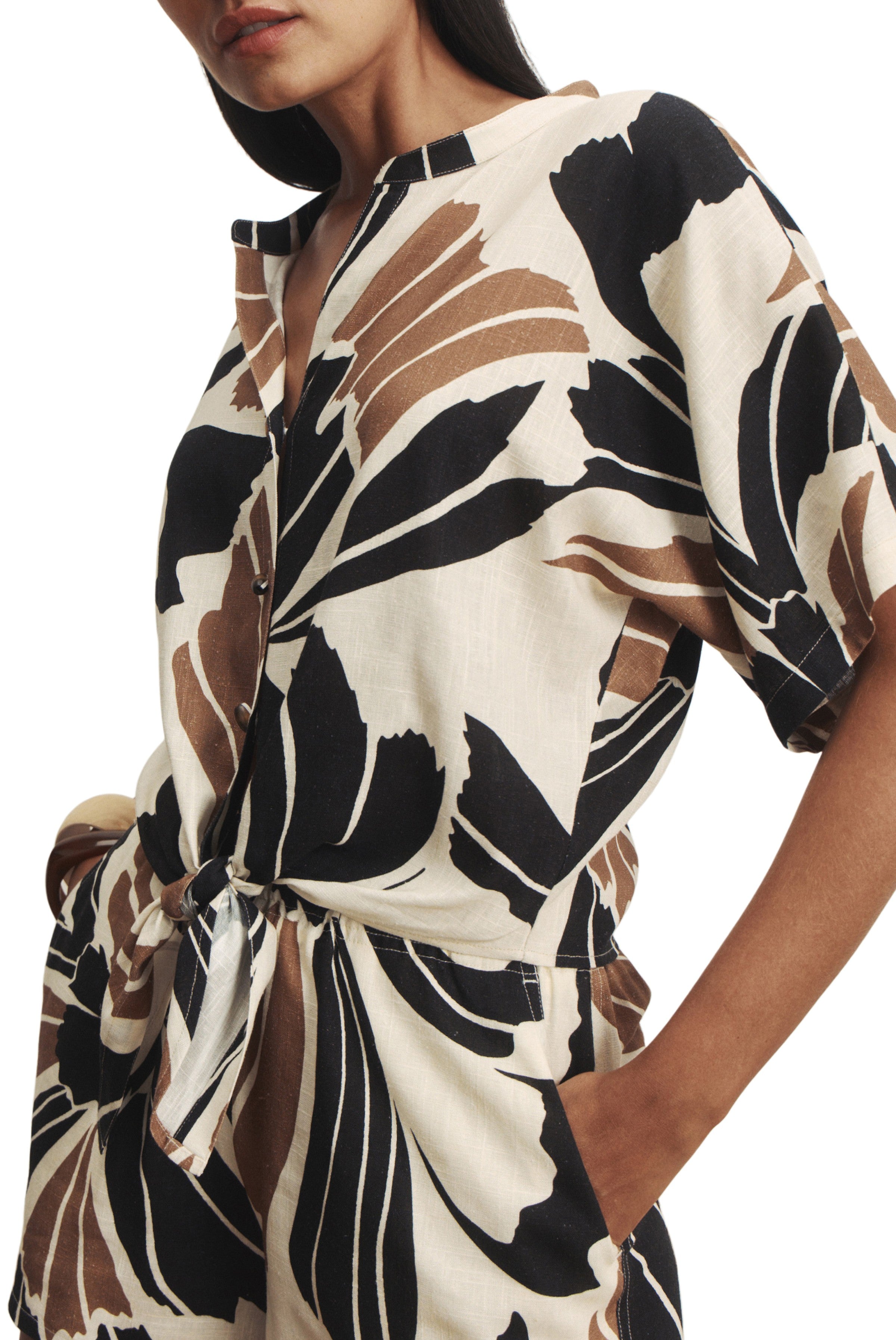 A woman models the Sanctuary Good Times Tie Shirt, a short-sleeve, button-up rayon linen romper with a beige, black, and brown abstract leaf print, relaxed fit, waist tie, and one hand casually in her pocket.