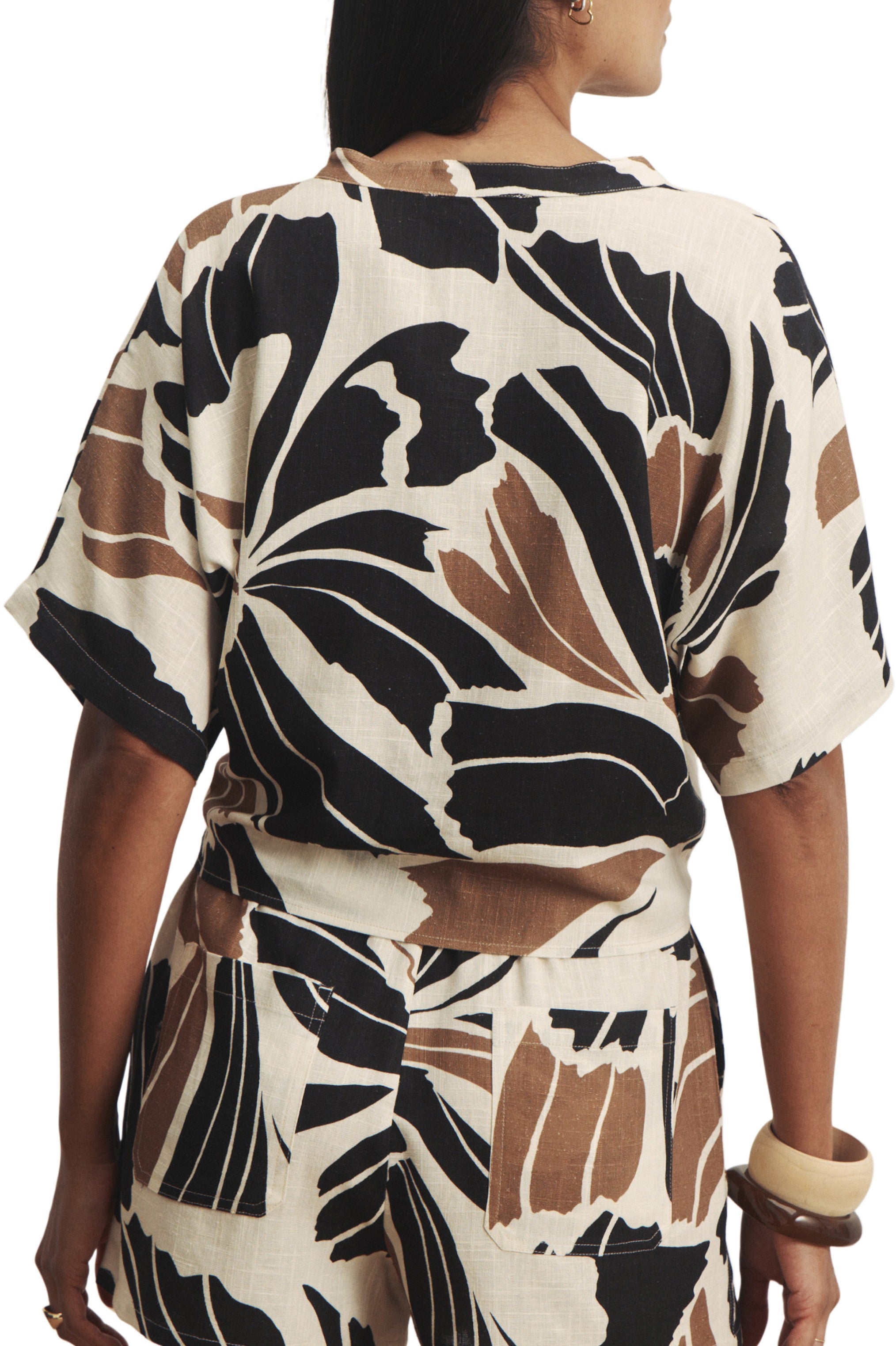 A woman seen from the back wears Sanctuary’s Good Times Tie Shirt in cream with a bold black and brown leaf print, paired with matching shorts and accessorized with a chunky bangle.