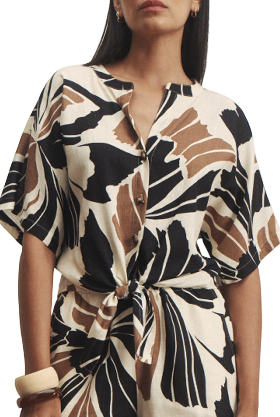 A woman wears the Sanctuary Good Times Tie Shirt in Black Multi, a relaxed fit dress with a bold abstract pattern, short sleeves, button-up front, and tie waist. She accessorizes with hoop earrings and a chunky bangle.