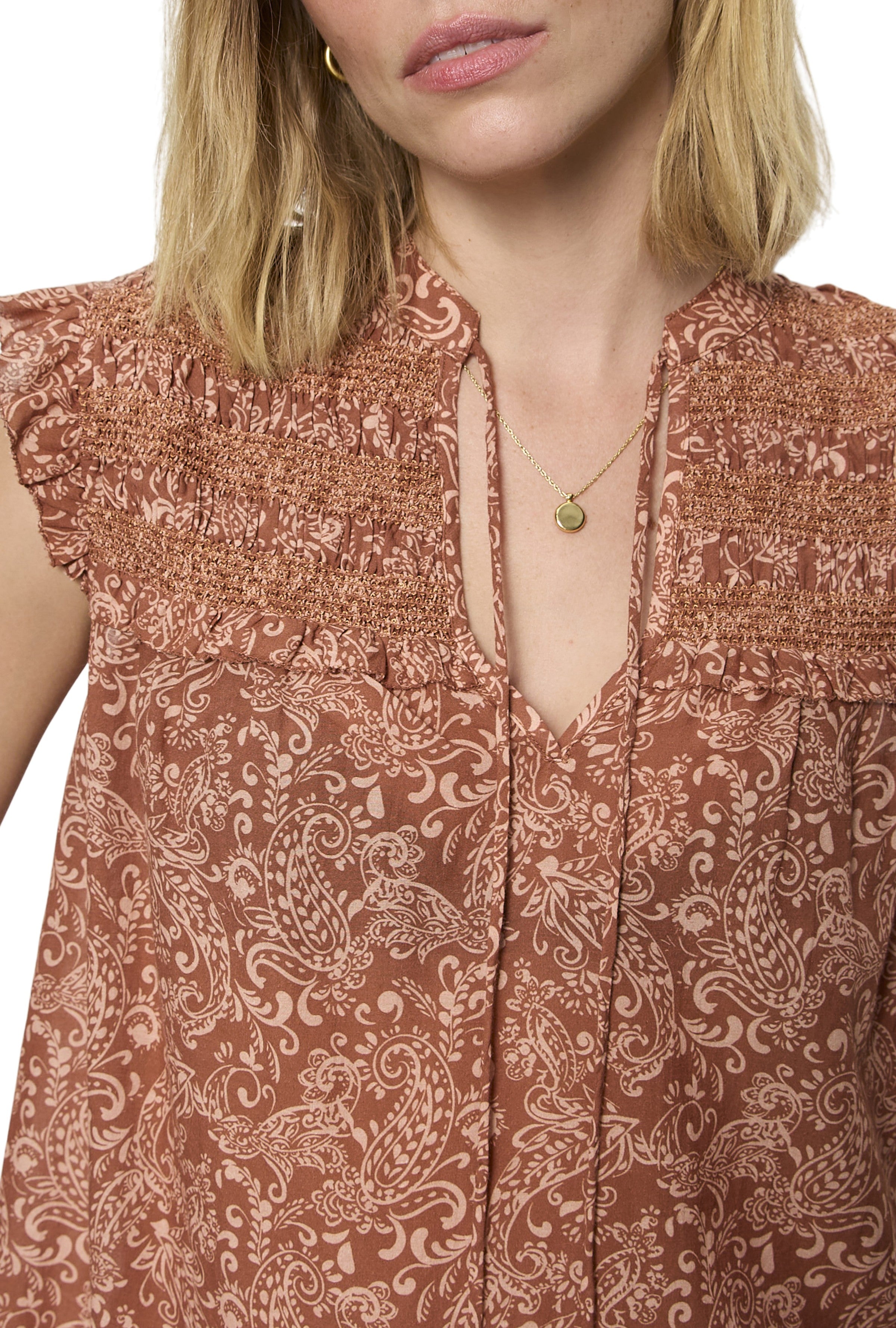 A woman wears the Sanctuary Free Feeling Smocked Blouse, featuring a brown and beige paisley pattern with smocked shoulders. She accessorizes with a gold necklace, her shoulder-length blonde hair partly covering her face.