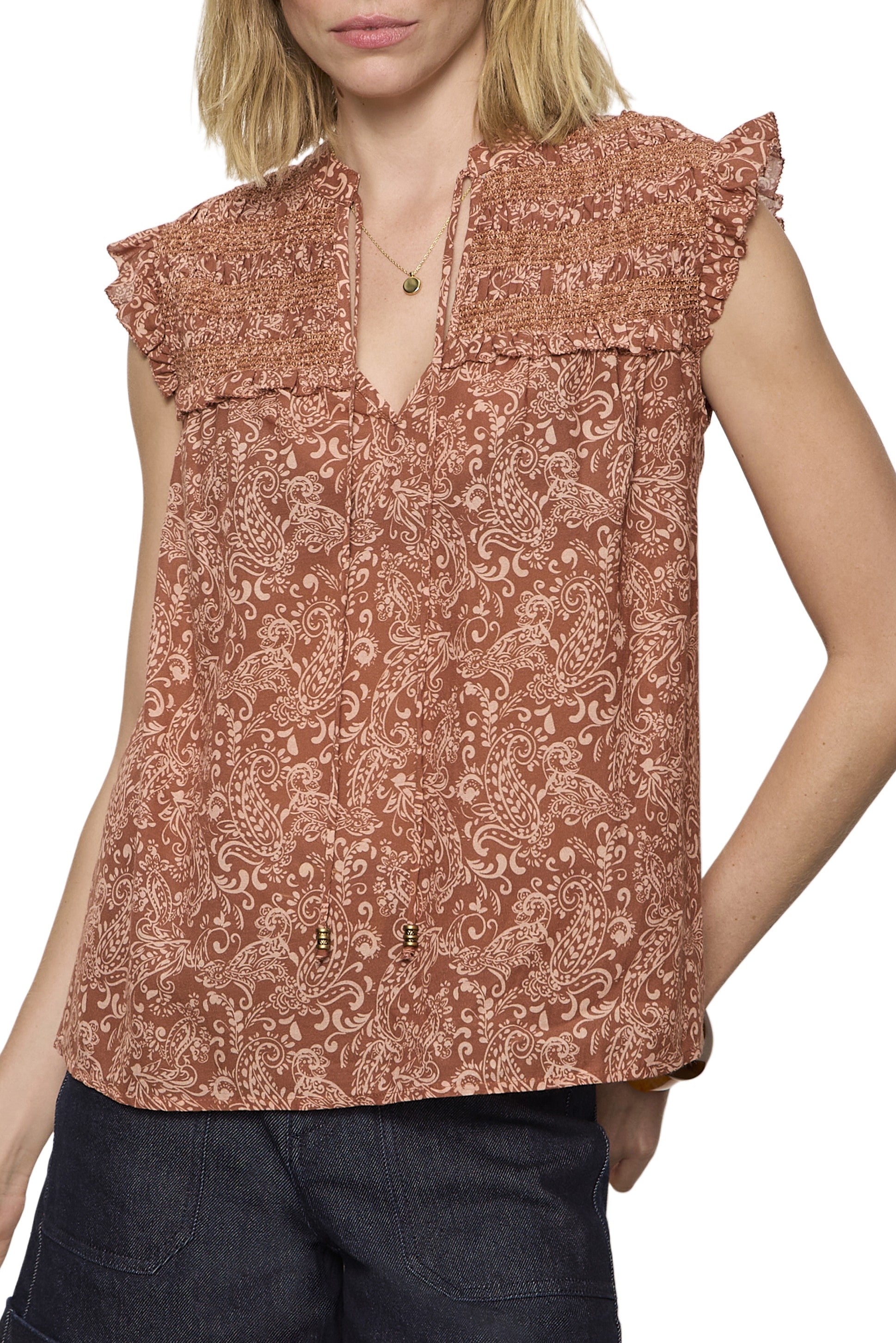 A woman wears the Sanctuary Free Feeling Smocked Blouse—a sleeveless, rust-colored top with a paisley print, ruffled shoulders, shirred yoke, relaxed fit, and slight V-neck—styled with a gold necklace and dark pants.