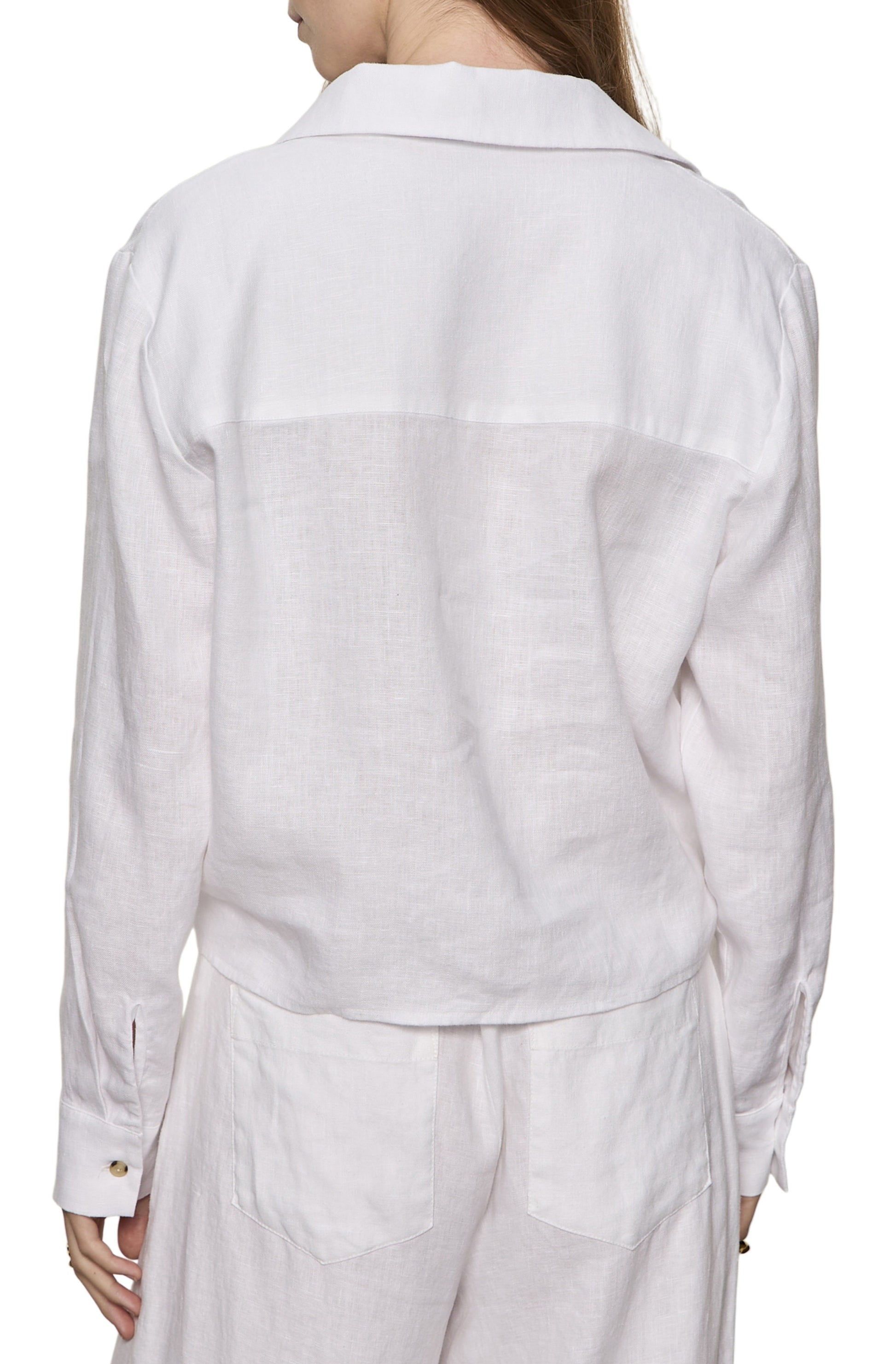 A woman is seen from behind wearing the Sanctuary More Love Linen Tie Top with long sleeves and button cuffs, paired with matching white linen pants featuring back pockets. Her straight hair drapes over one shoulder against a clean white background.