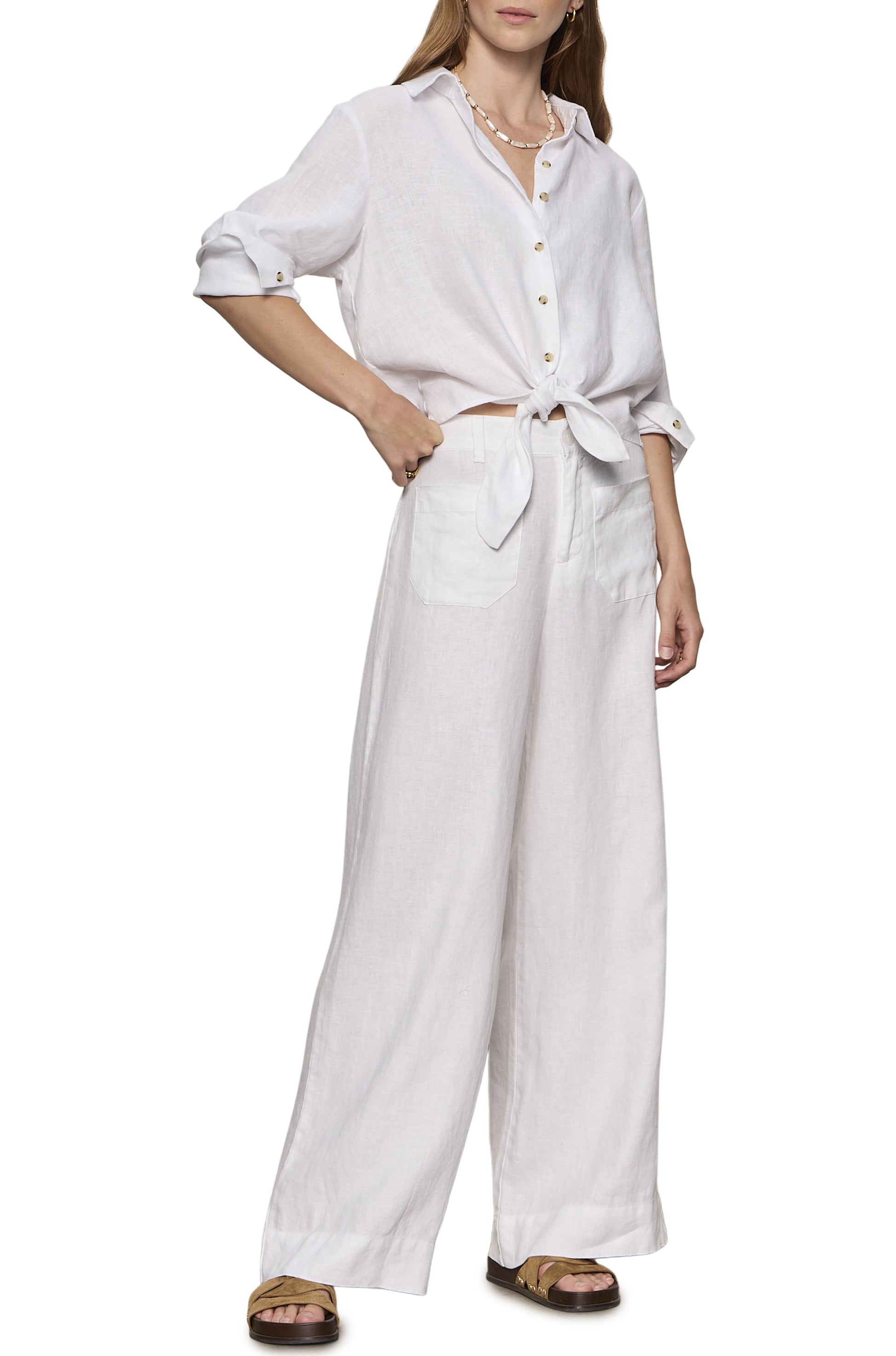 A woman wears the Sanctuary More Love Linen Tie Top with wide-leg white pants, a necklace, and sandals. Her right hand is on her hip.