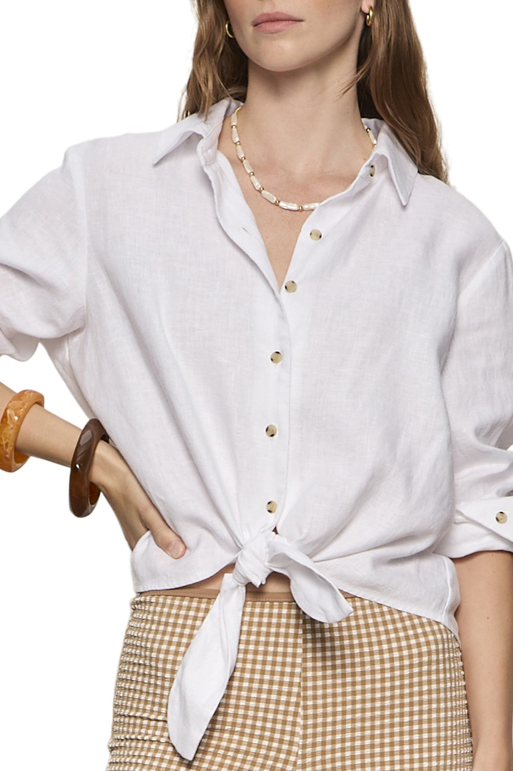 A woman in the Sanctuary More Love Linen Tie Top in white, tied at the waist, stands confidently with one hand on her hip, styled with brown plaid pants, a shell necklace, and brown bangles.