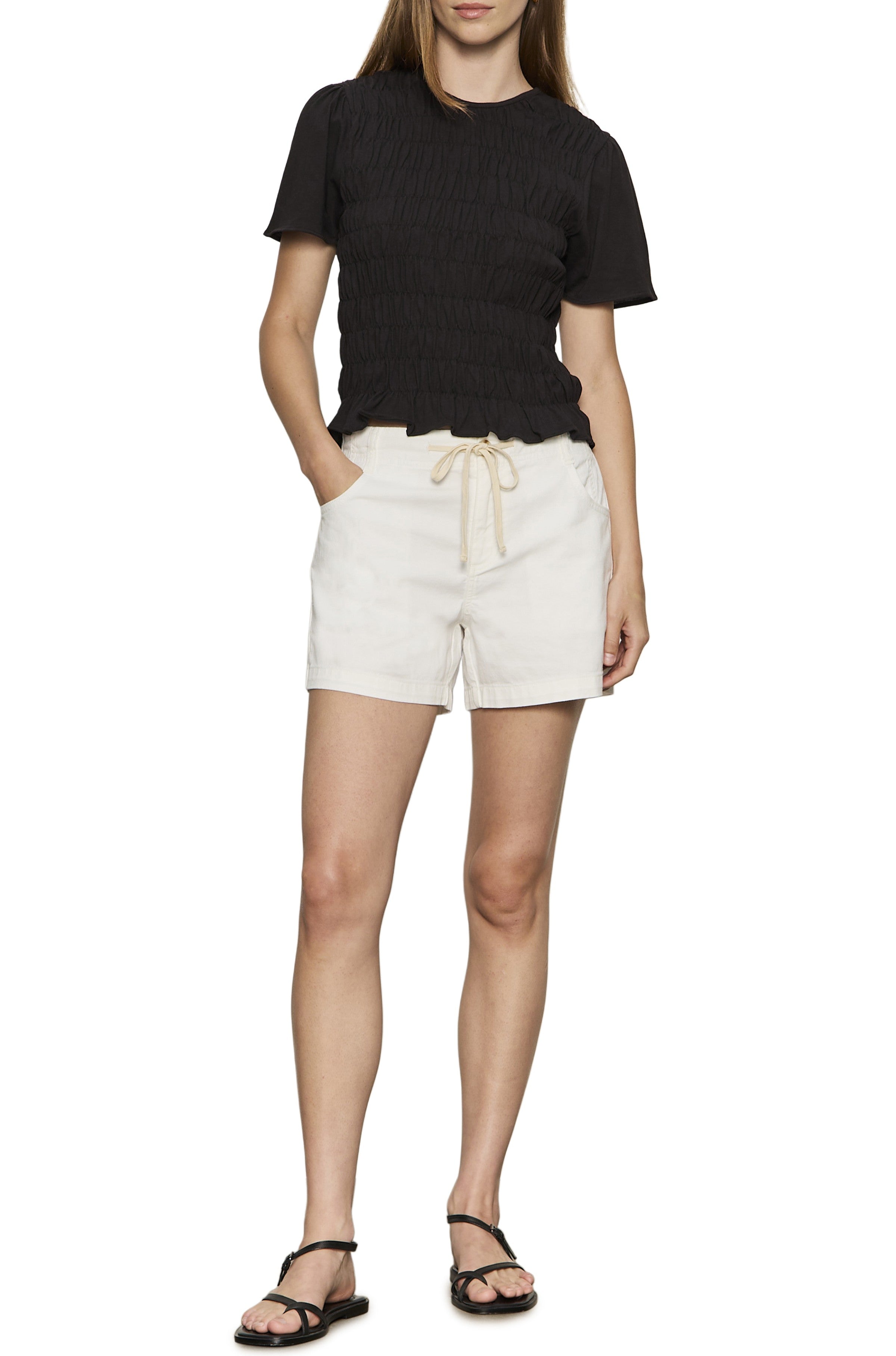 A person stands with one hand in their pocket, wearing a black short-sleeve shirt, black sandals, and Sanctuary's Idyll Tie Short—off-white drawstring shorts with an elasticated waistband.