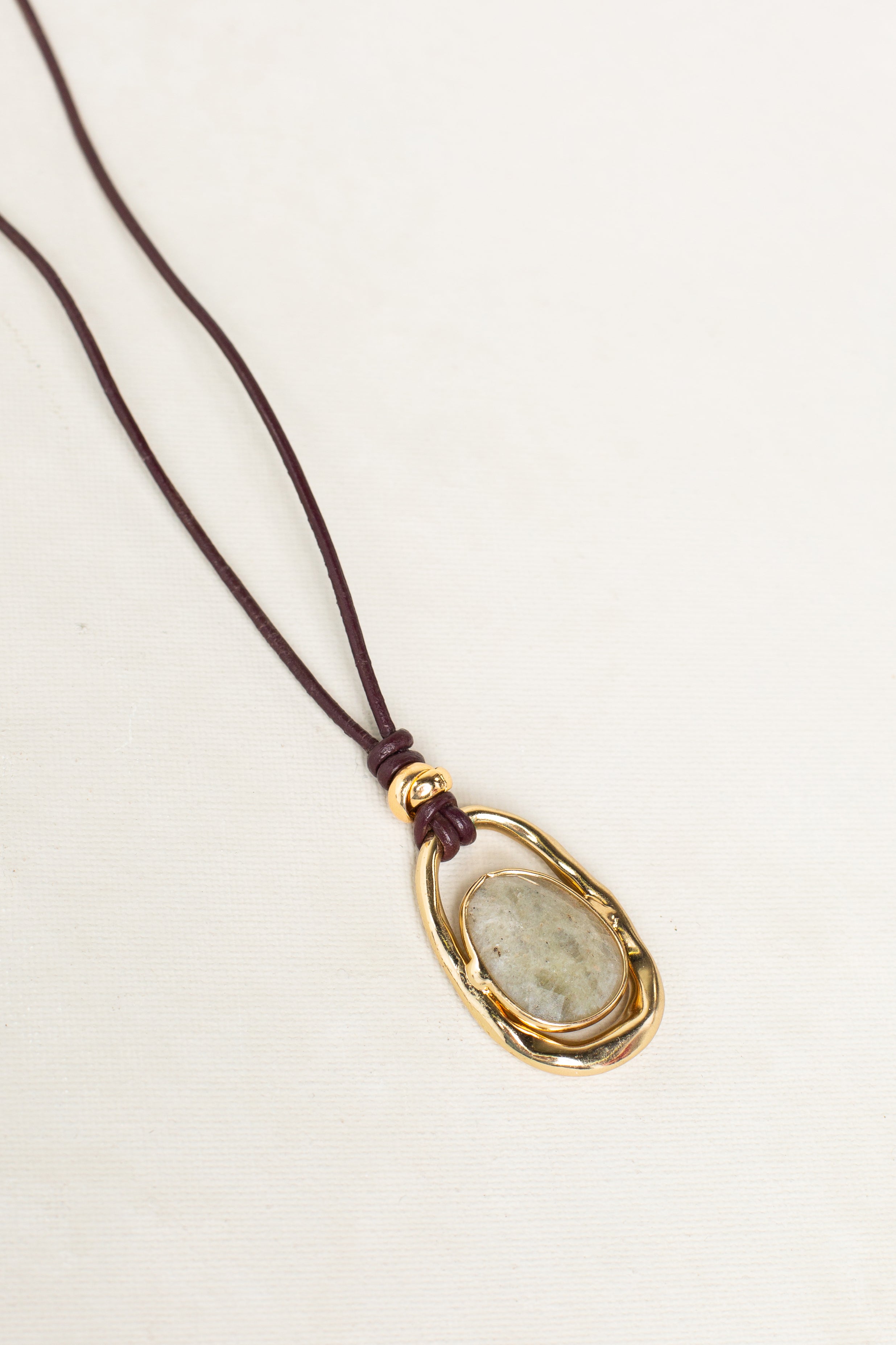 The Isla x Tate Adjustable Leather Cord Pendant Necklace features a gray oval gemstone set in a gold-tone frame on a dark brown leather cord, shown on a light background. Ideal for statement jewelry lovers.