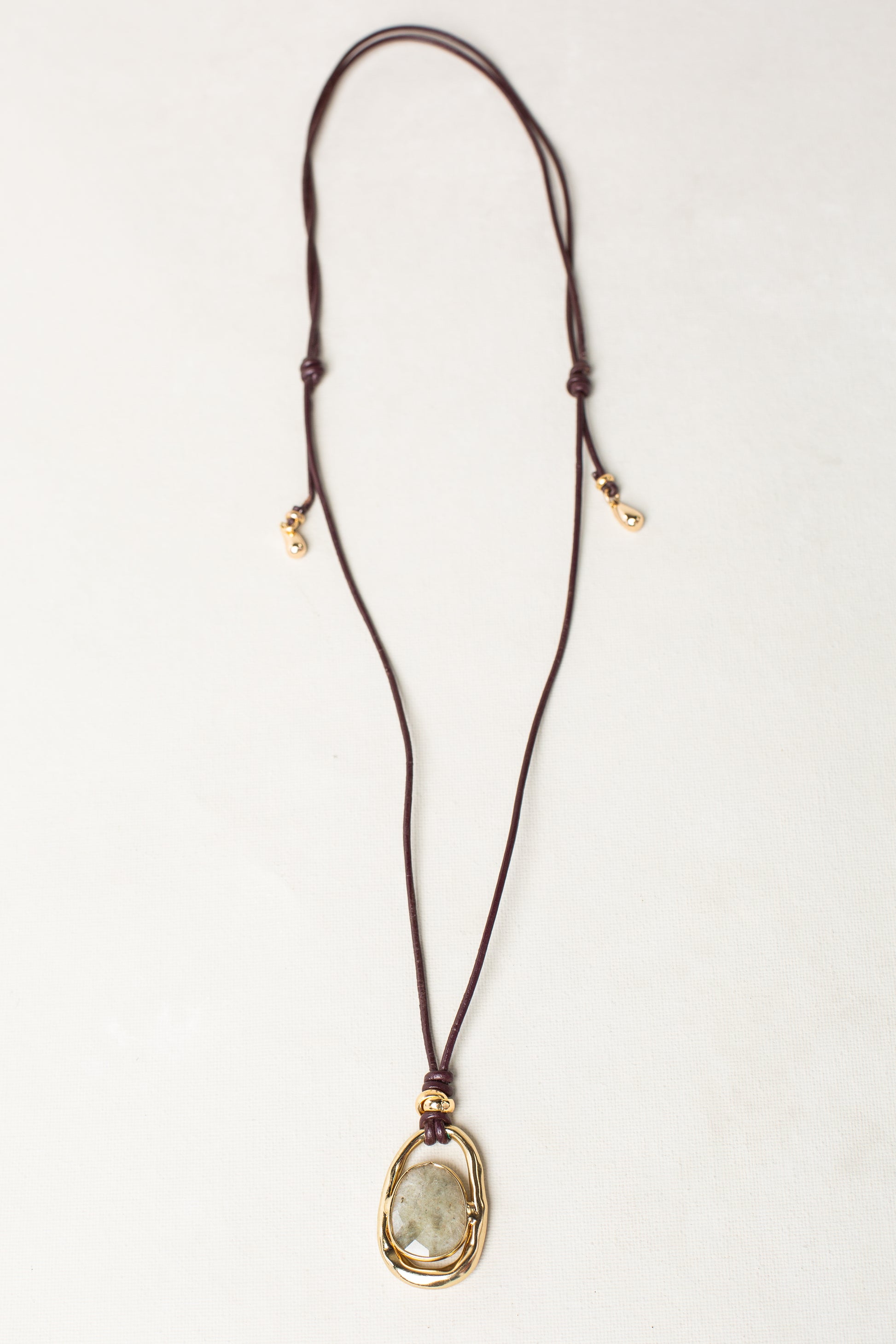 The Isla x Tate Adjustable Leather Cord Pendant Necklace features a gold wire-wrapped, oval semi-precious stone on a brown leather cord—a striking statement piece against any light background.