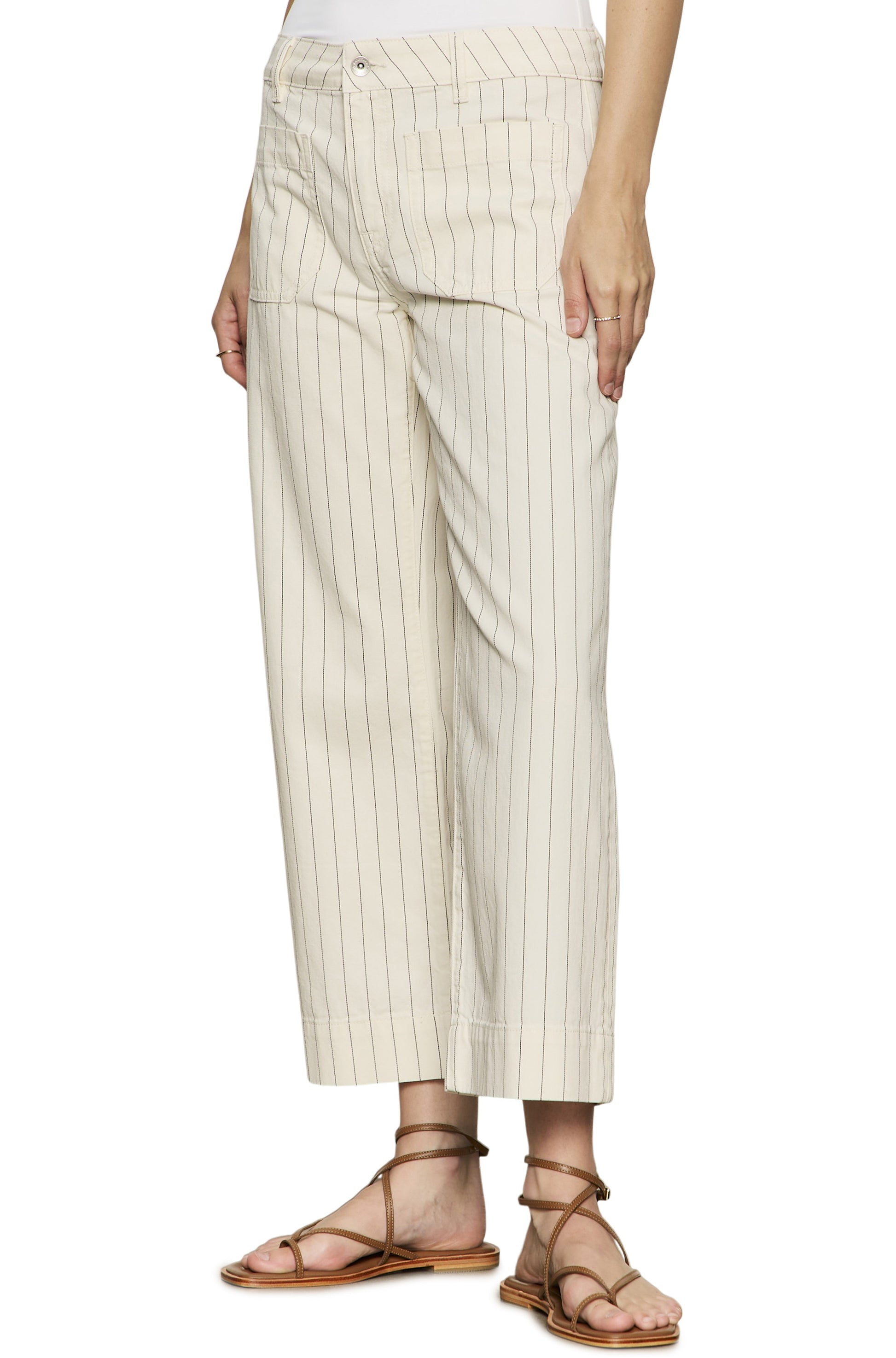A person wears Sanctuary's The Marine Bleached White Pinstripe cropped wide leg pants with a relaxed fit and cream pinstripes, crafted from cotton, paired with brown strappy flat sandals. Only the upper body is partially visible.