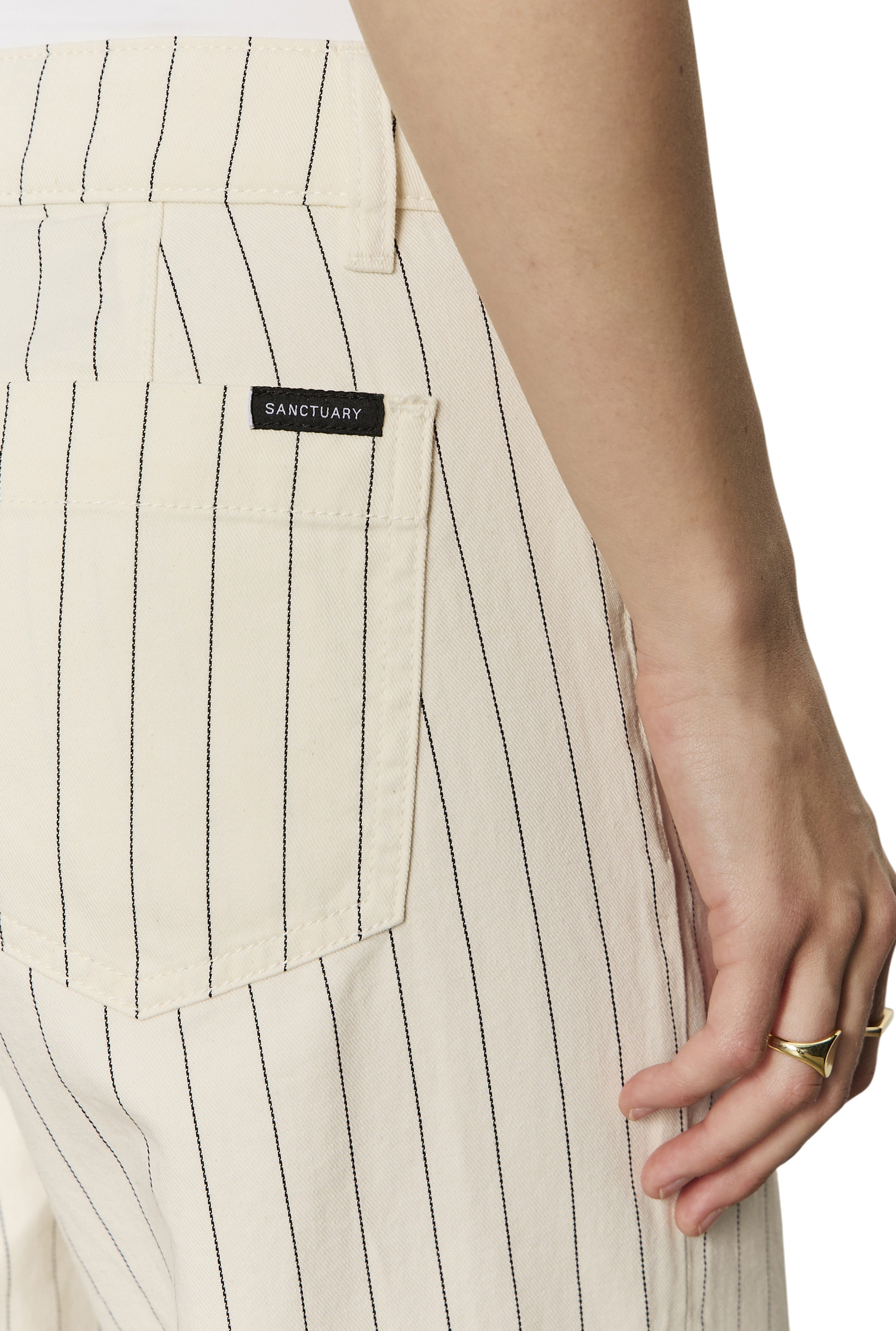 A close-up of a person wearing Sanctuary's The Marine Bleached White Pinstripe cotton cropped wide leg pants with thin black stripes, a rear pocket, and a Sanctuary label. Their gold-ringed hand rests at their side.