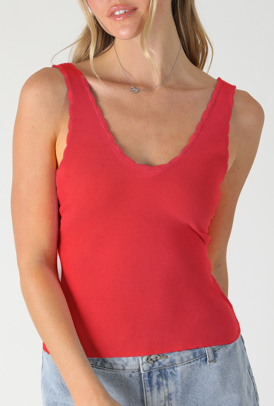 A woman wears the Niki Biki Scallop Edge Tank Top in hot pink, styled sleeveless with a V-neck and scalloped edges, paired with light blue jeans, shown waist up against a plain light background.