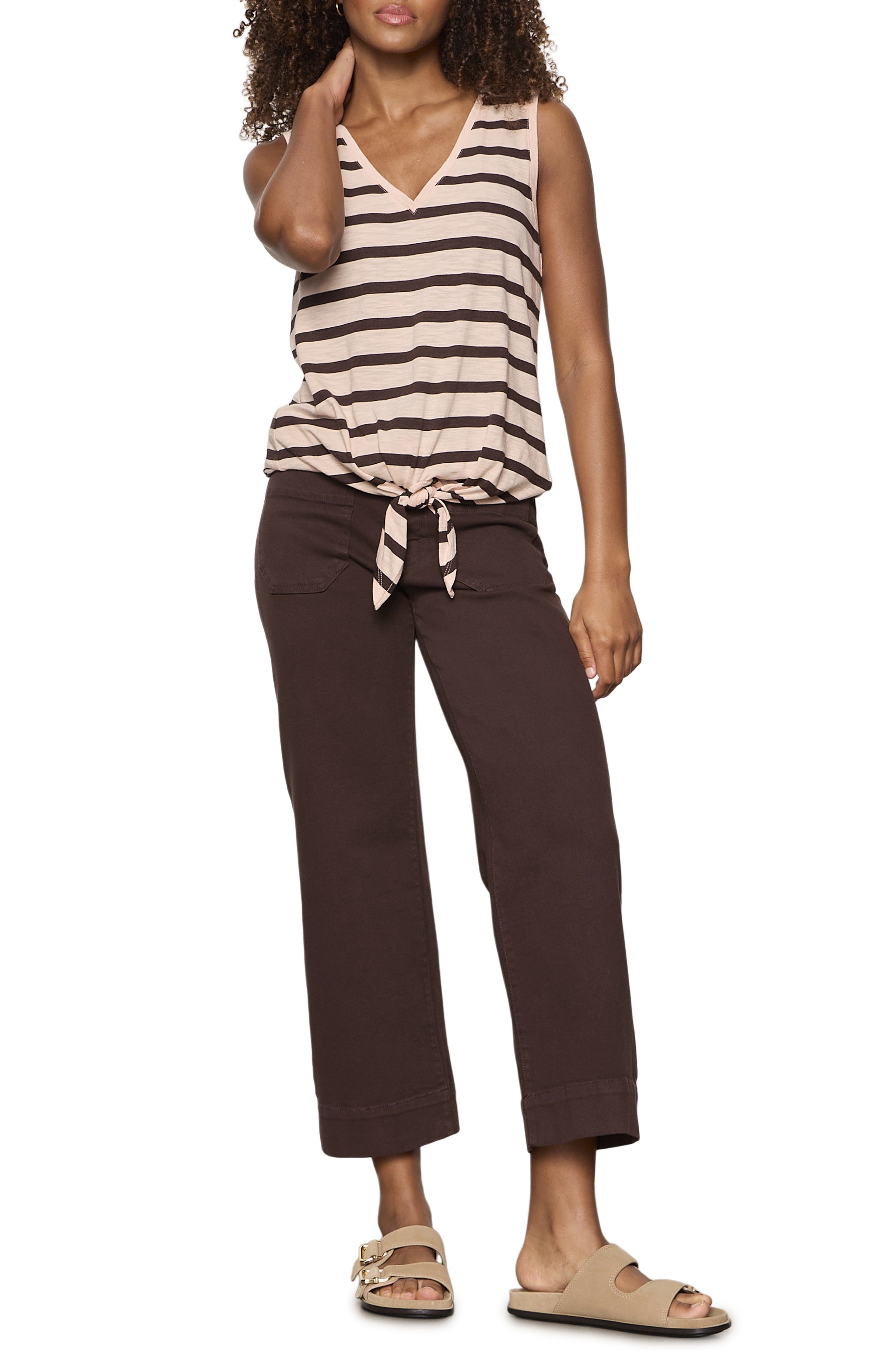 A woman wears the Sanctuary V Neck Tie Tank—sleeveless with beige and black stripes, a front tie, and relaxed cotton rayon fabric—paired with dark brown wide-leg pants and beige slides. She poses with one hand in her hair.