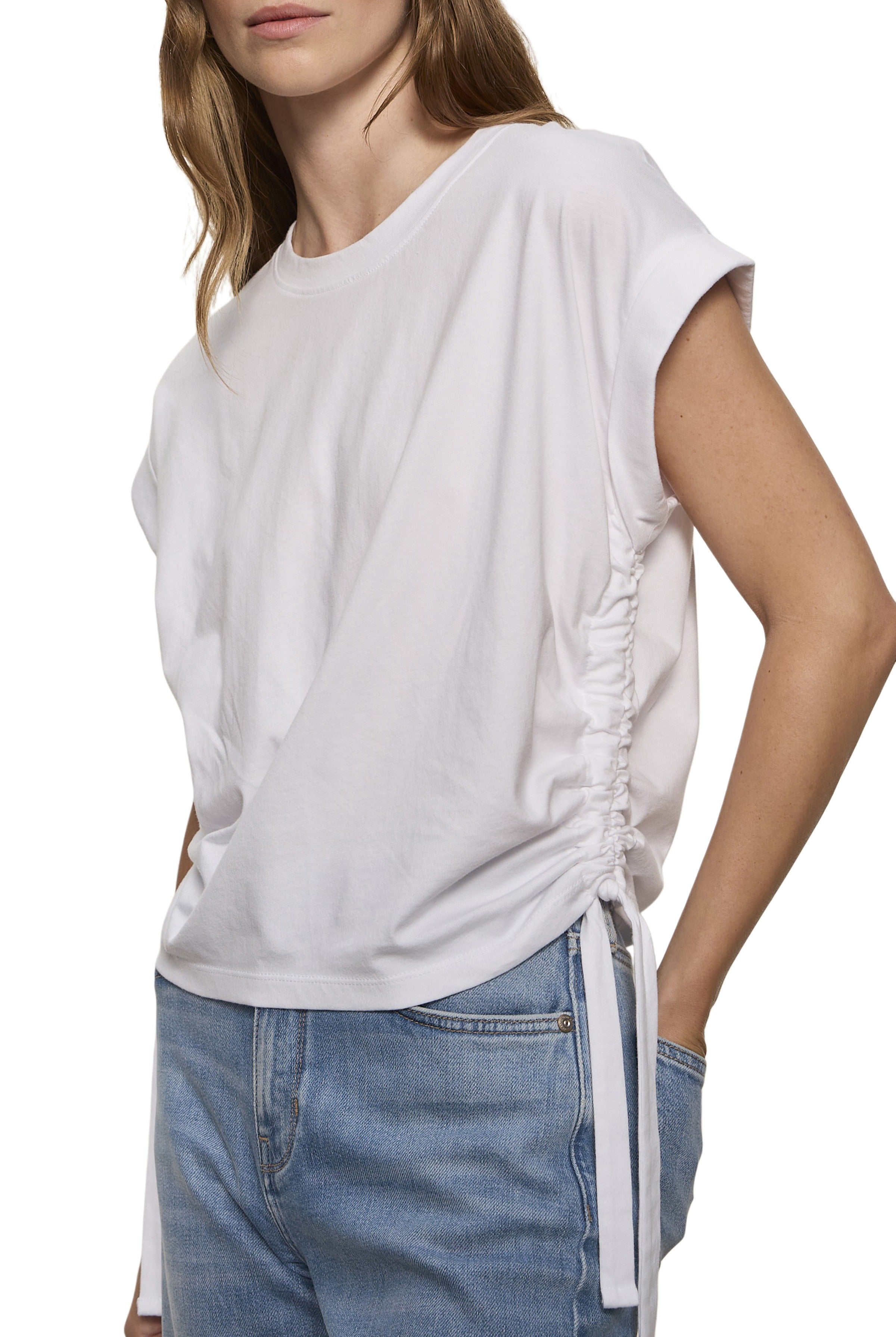 A woman wears the Sanctuary All Day Ruched Tee in white, featuring adjustable side ruching and a tie, paired with light blue jeans. The photo is cropped from her shoulders to mid-thigh.