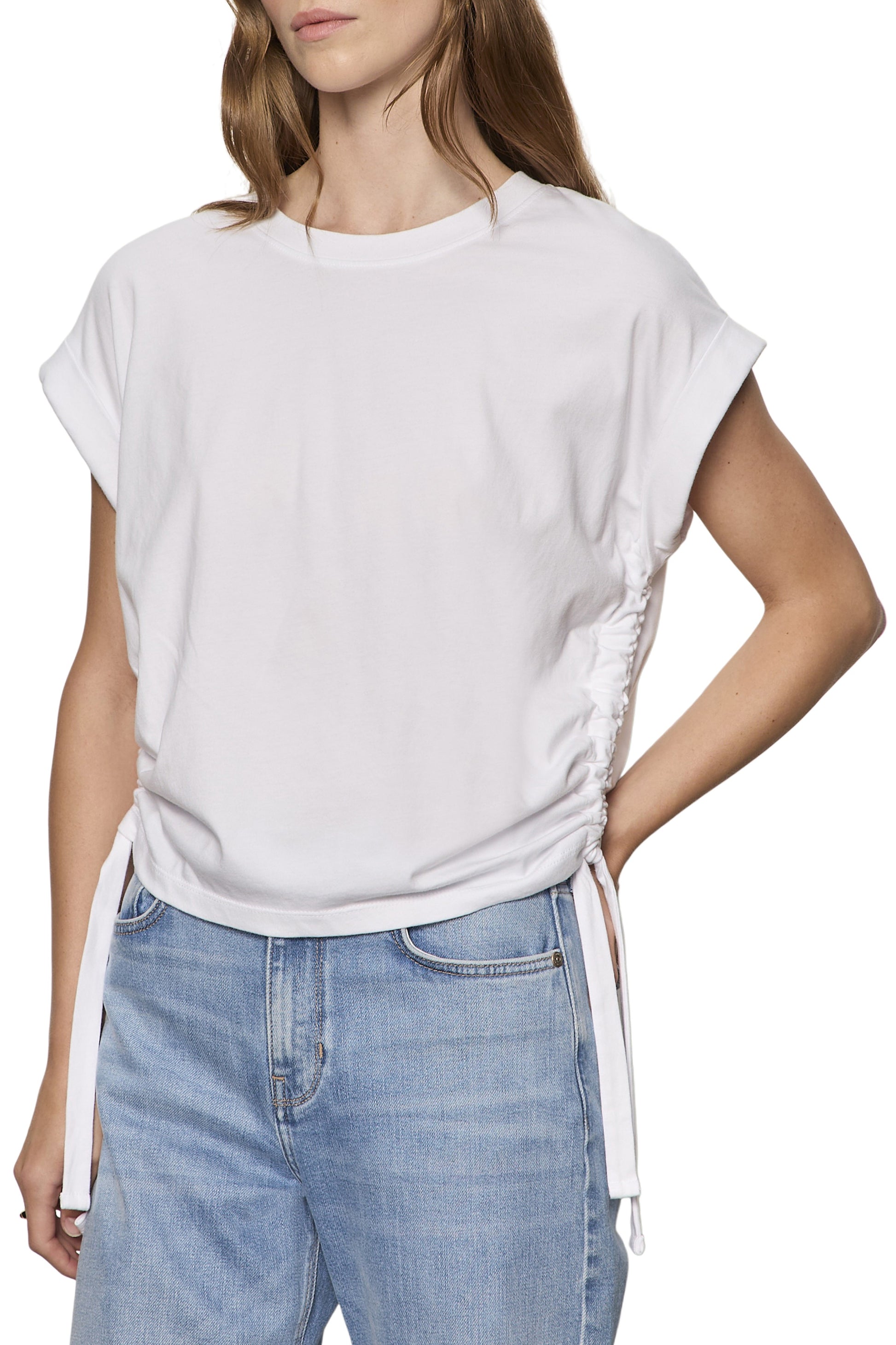 A woman models the Sanctuary All Day Ruched Tee in white, featuring adjustable side ruching and ties, paired with light blue high-waisted jeans. She stands relaxed with one hand on her hip, her hair down.