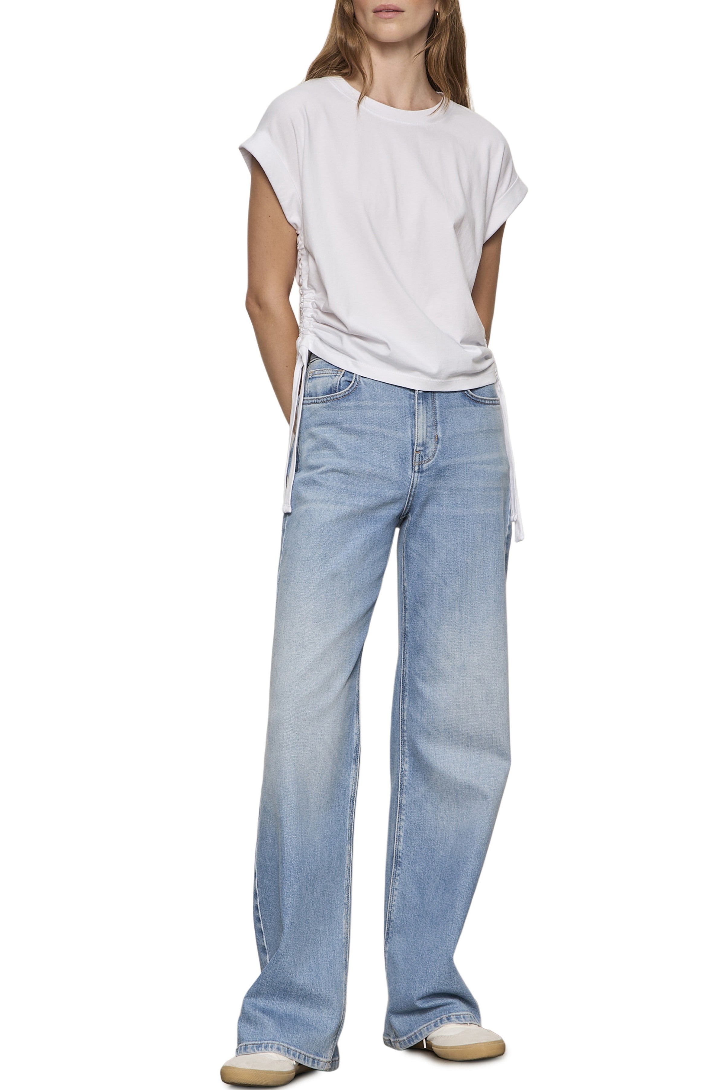 Wearing the Sanctuary All Day Ruched Tee in white with light blue high-waisted wide-leg jeans and beige shoes, a person stands hands behind their back against a white background.