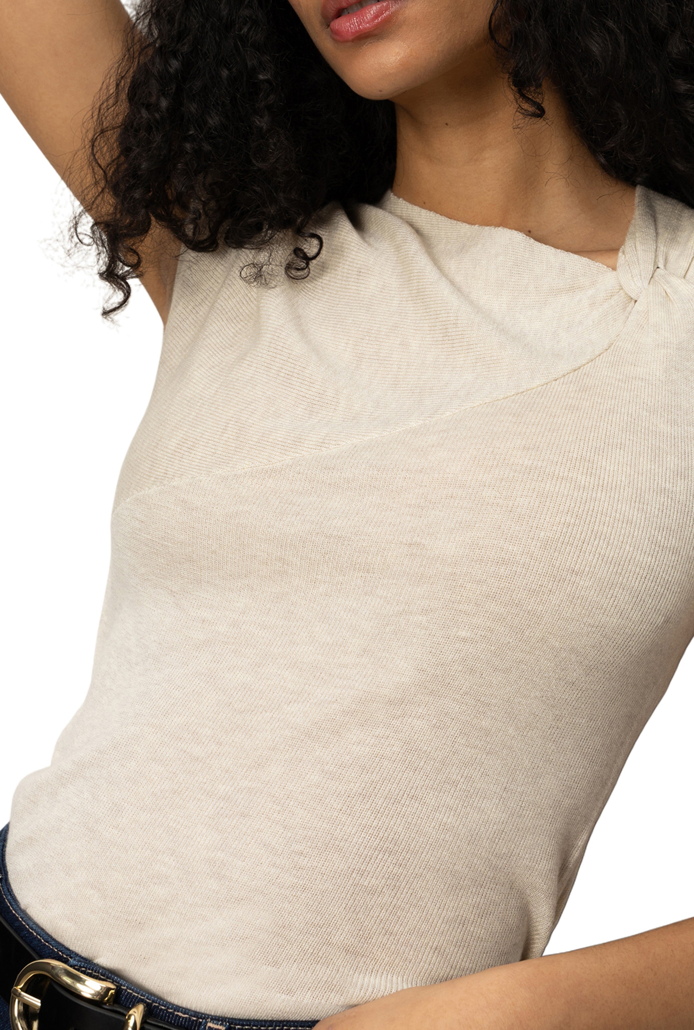 A woman with curly dark hair wears Sanctuary's Asymm Shoulder Twist Top—a fitted, cream ribbed knit sleeveless top with a unique twist neckline—paired with dark blue jeans and a gold-buckle belt.